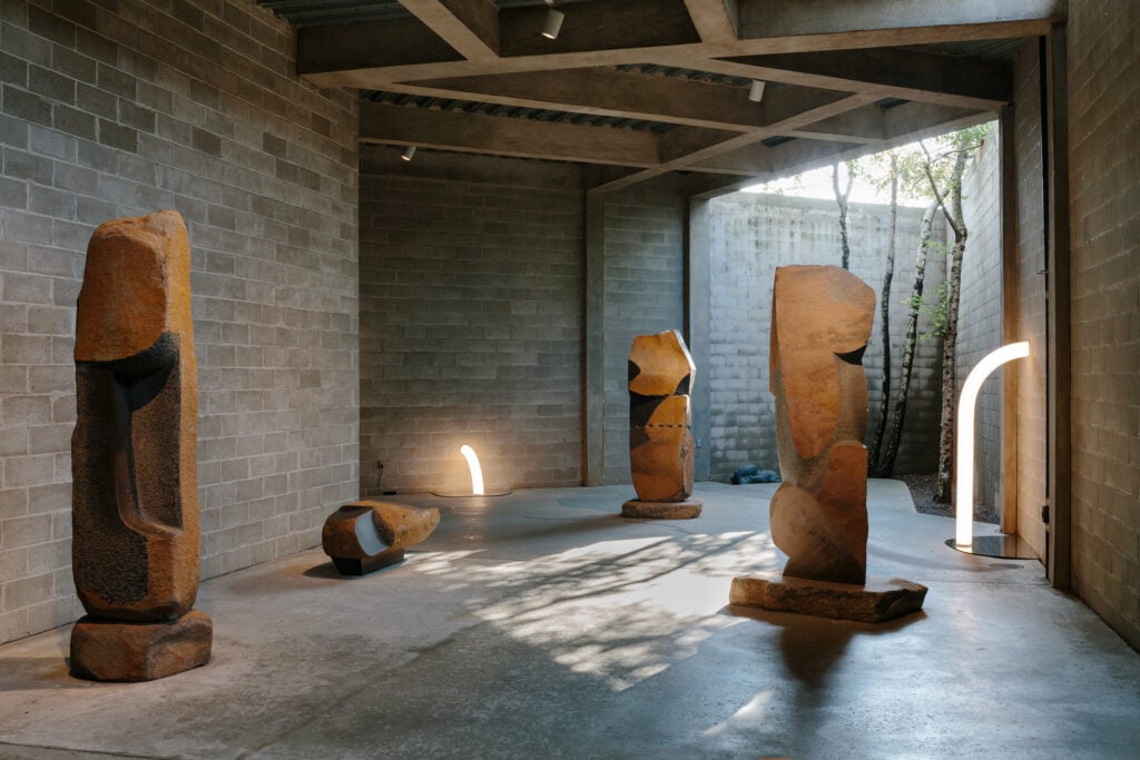 Light sculptures and stone sculptures sitting on the floor of a concrete open air gallery with trees growing in the corner. 