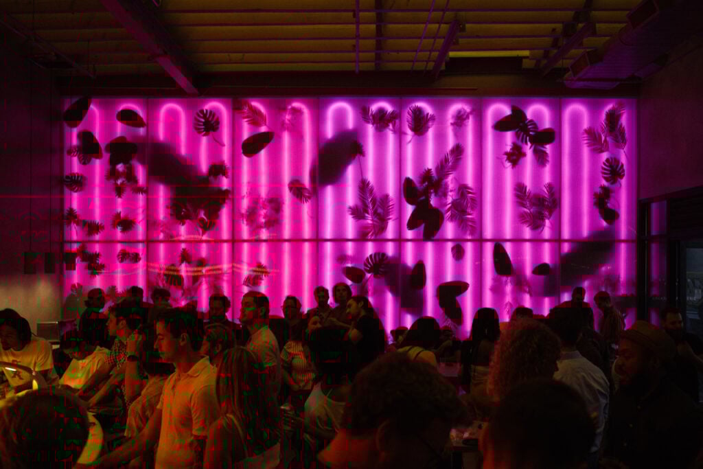 Interior of a contemporary bar filled with people in front of a large backlit wall with plants showing through.