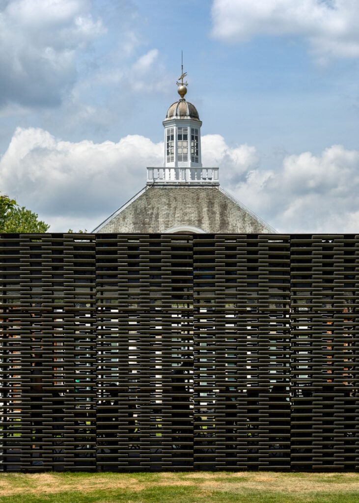 the roof of an old-fashioned building appears over the serpentine pavilion wall