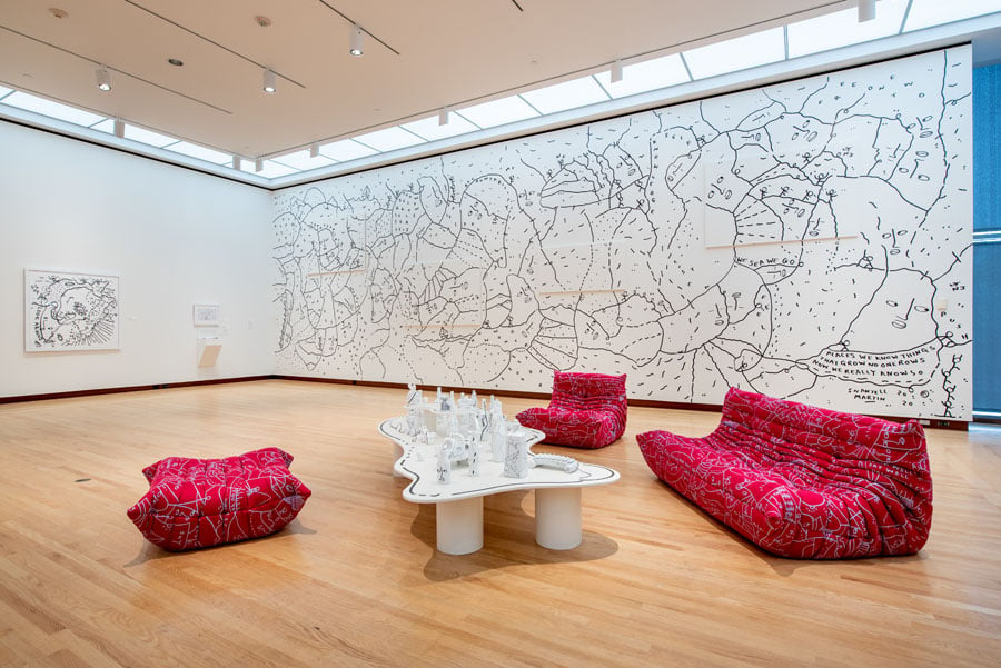 Black line drawings on white walls with red cushion lounge chairs and white table, all by Shantell Martin.