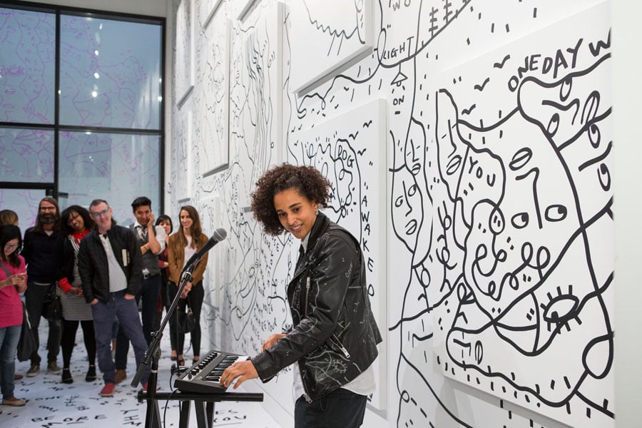 Shantell Martin playing keyboard next to a wall and floor covered by her black and white line drawings.