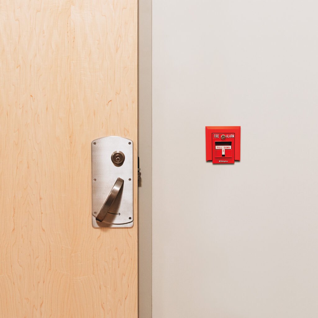 A door knob with simple details in stainless steel placed closed to the fire alarm on the wall next to it. 