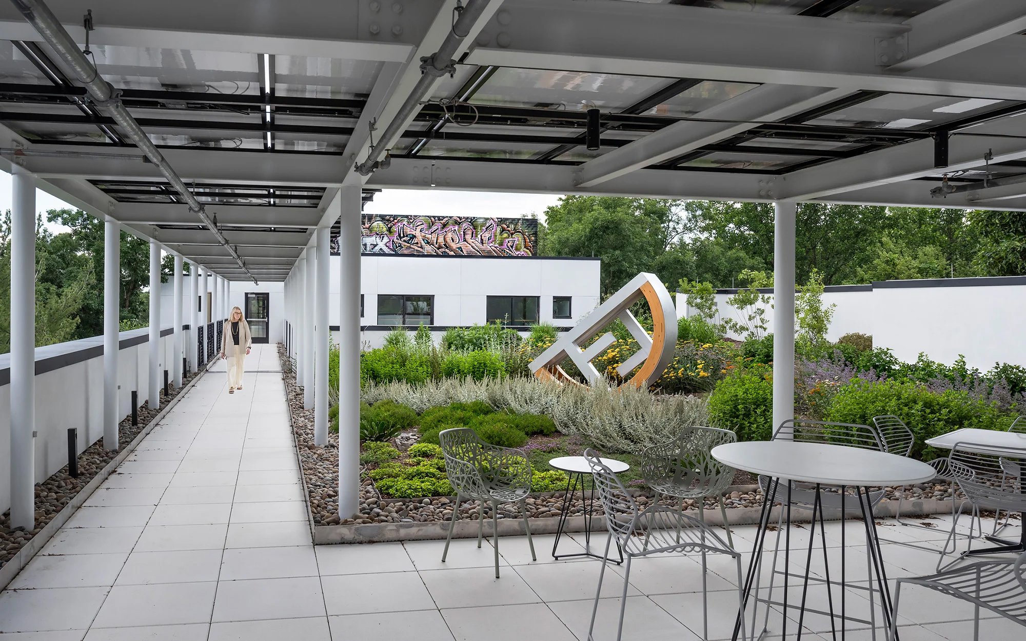 Woman walking on the covered rooftop of the HMTX building which is covered with some furniture and green herbs and plants.