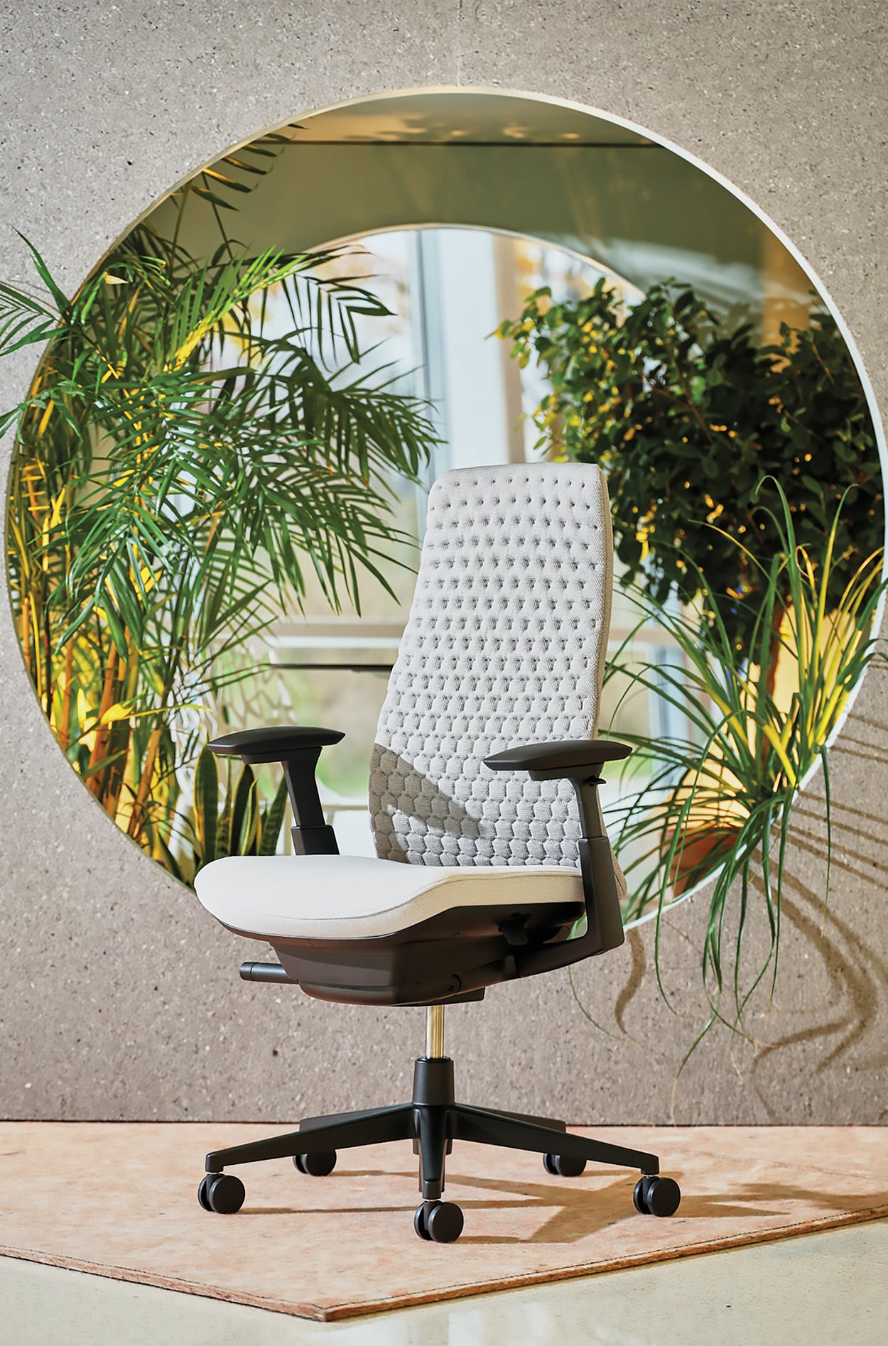 A white woven fern chair kept in the centre of a circle cut out wall with a lot of plants in the background. 
