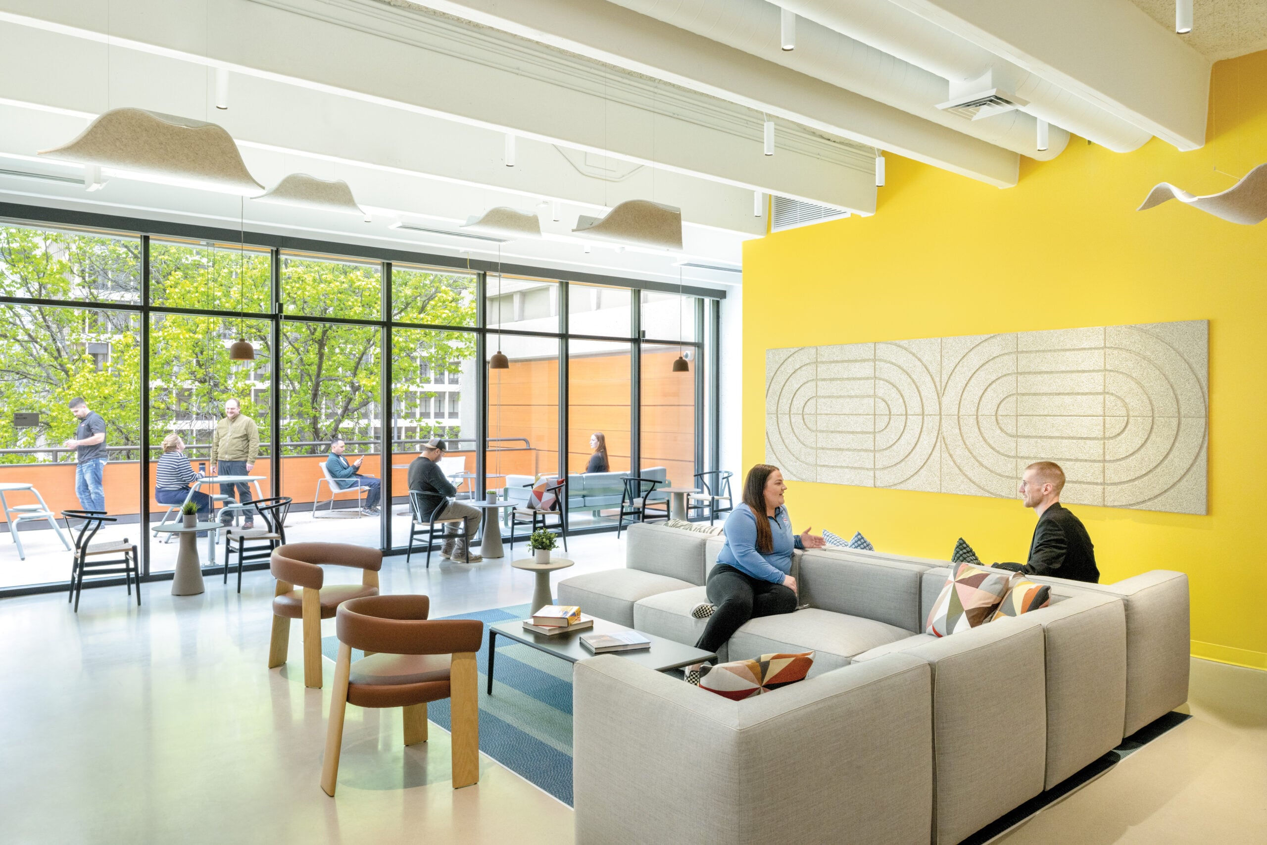 A bright, modern lounge area inside the Bateson Building featuring a yellow wall with acoustic art, grey sectional sofas, and white acoustic ceiling baffles. Large windows open onto an outdoor terrace where people are working at tables.
