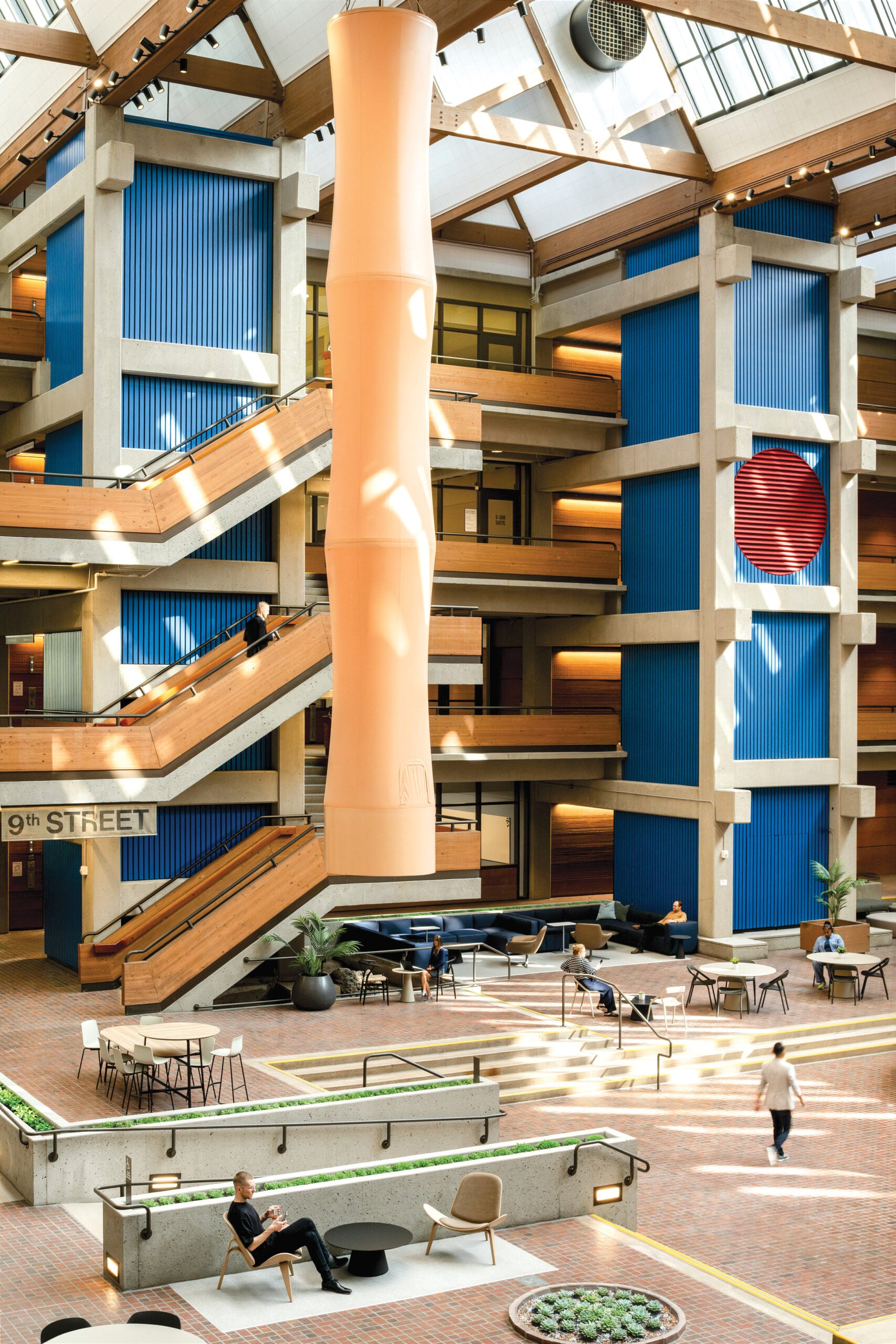 A soaring interior atrium with a large vertical orange tube (fan) hanging in the centre, wooden staircases leading to multiple floors and a ground floor seating area.
