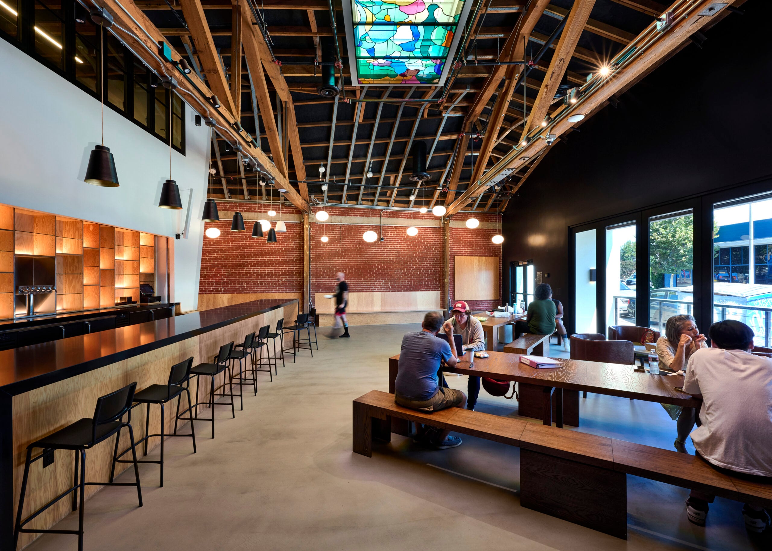 A spacious, modern theatre lobby featuring a high vaulted ceiling with exposed wooden trusses and a long, colorful stained-glass skylight. A bar with stools lines the right side, while people gather at communal wooden tables and benches on the left.