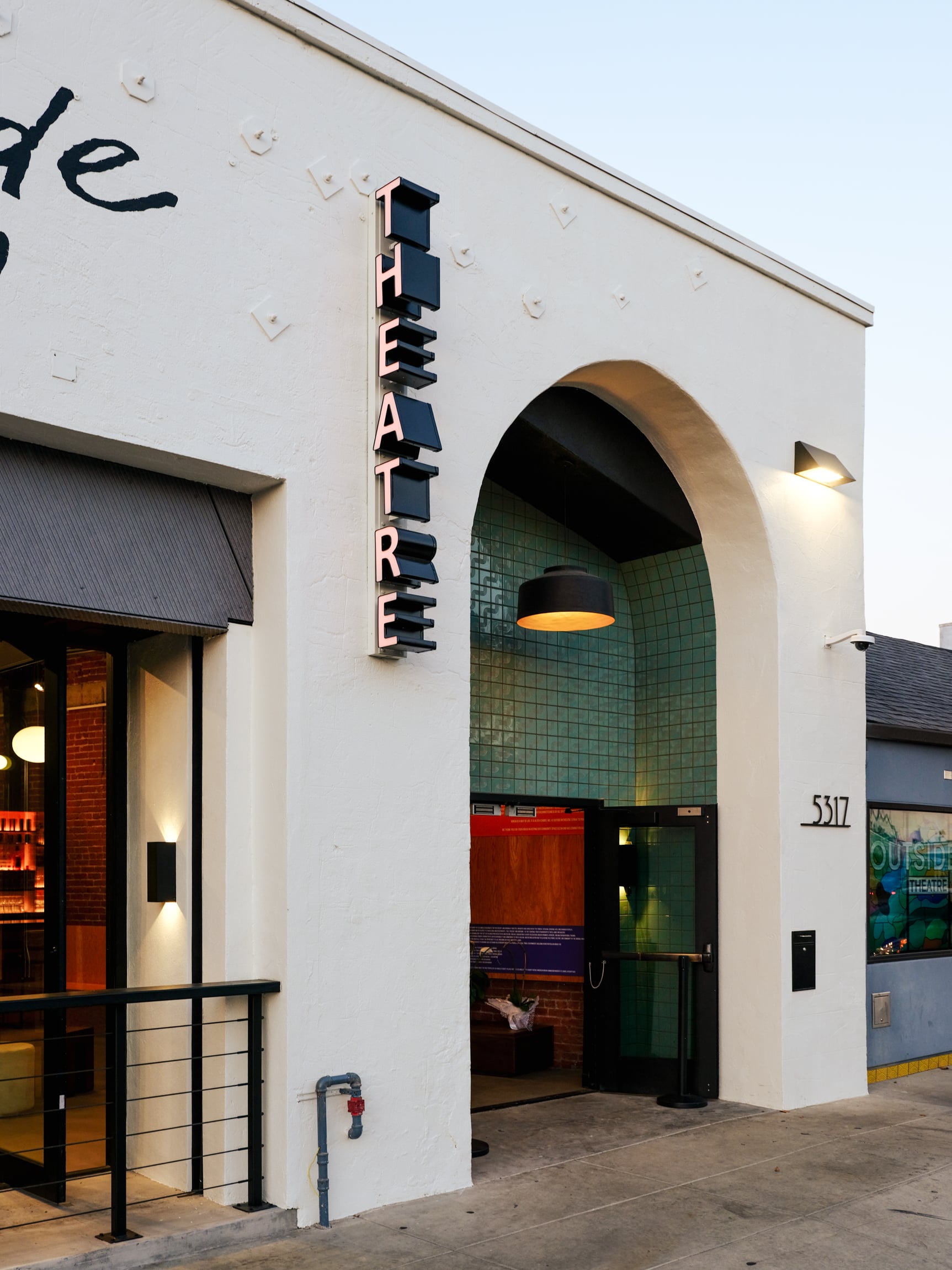 The exterior of a white stucco building with a large arched entryway. A vertical marquee sign attached to the building reads 'THEATRE' in bold, illuminated letters.