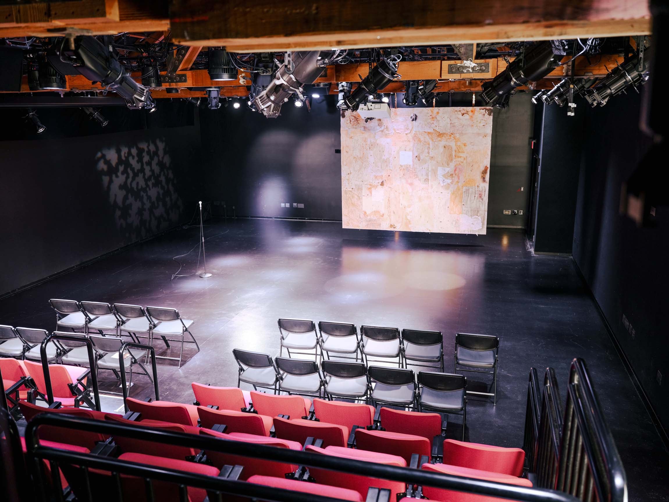 A view inside the darkened theatre focusing on the tiered seating bank featuring red upholstered chairs. A screen is visible on the far left wall, and the industrial ceiling grid is exposed above.