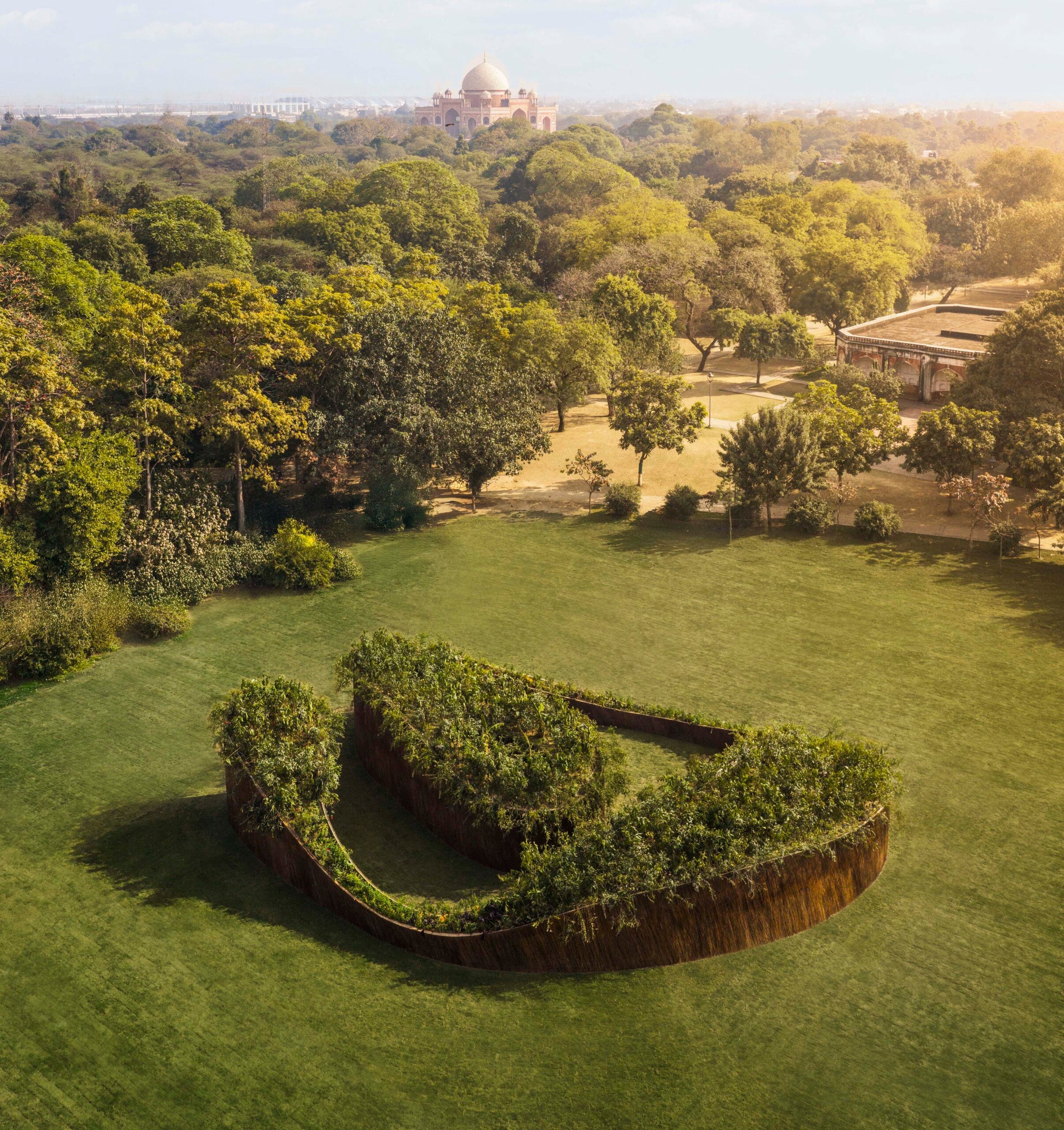 An elevated top-down shot showing the pavilion’s circular, "O" shaped layout. The structure is topped with lush green vegetation and sits in a large, open grassy field surrounded by mature trees.