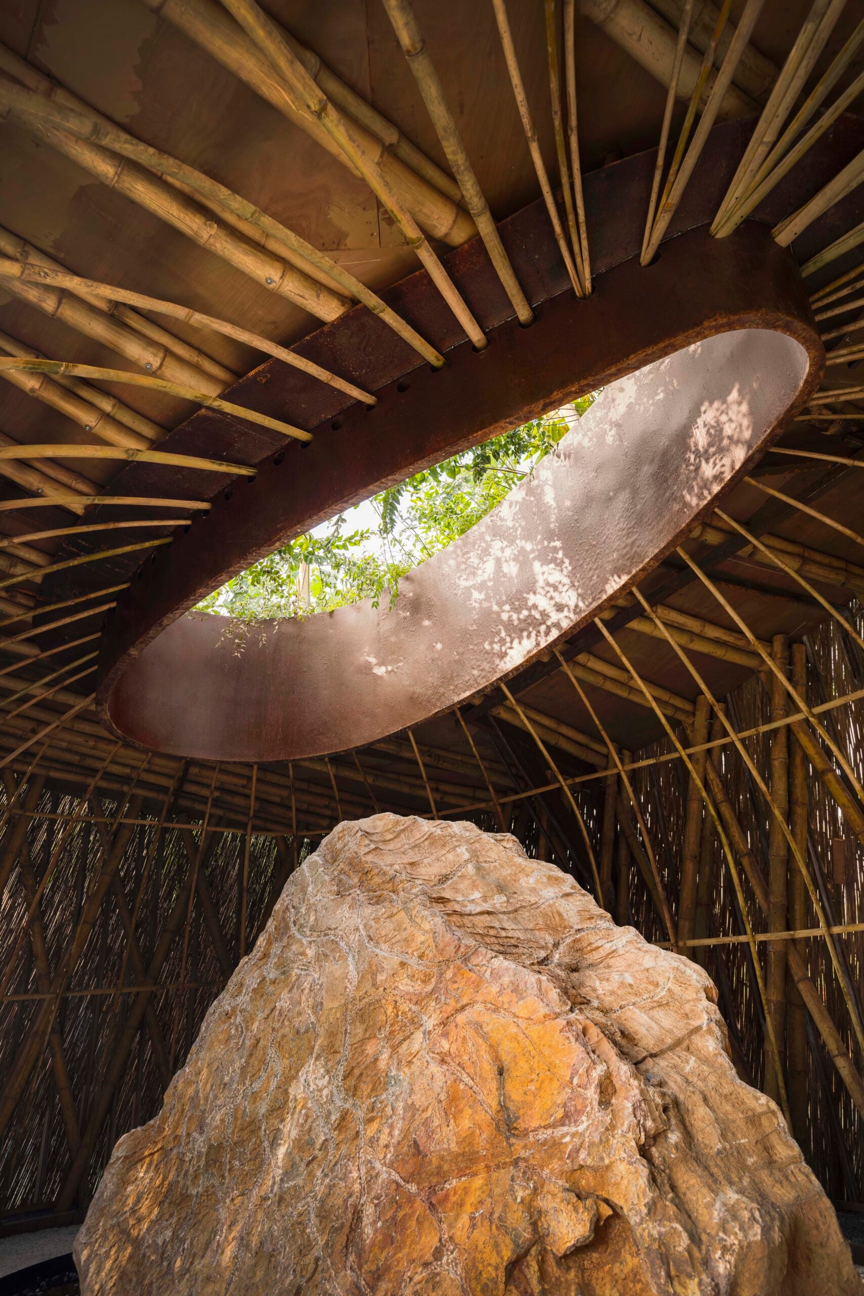 An interior shot looking directly up at a circular opening in the woven bamboo roof. Sunlight streams through the aperture, illuminating the intricate wooden lattice and the large standing stone beneath it.