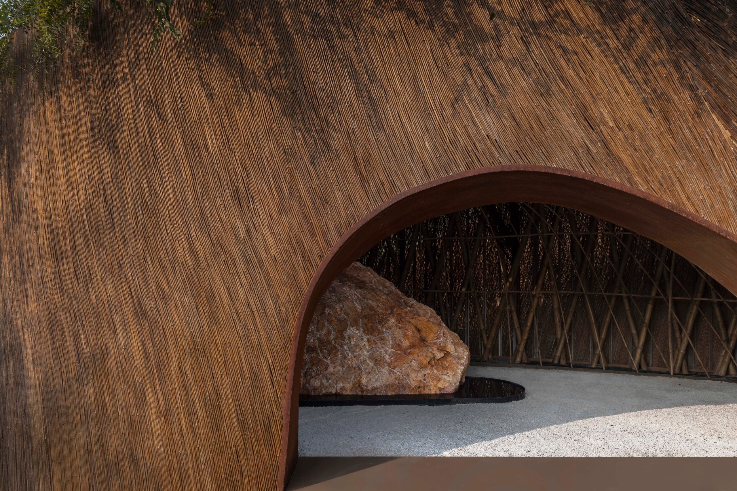 A close-up of a low, rounded archway leading into the pavilion. The thick, reddish-brown textured walls frame a view of the gravel floor and the central boulder inside.