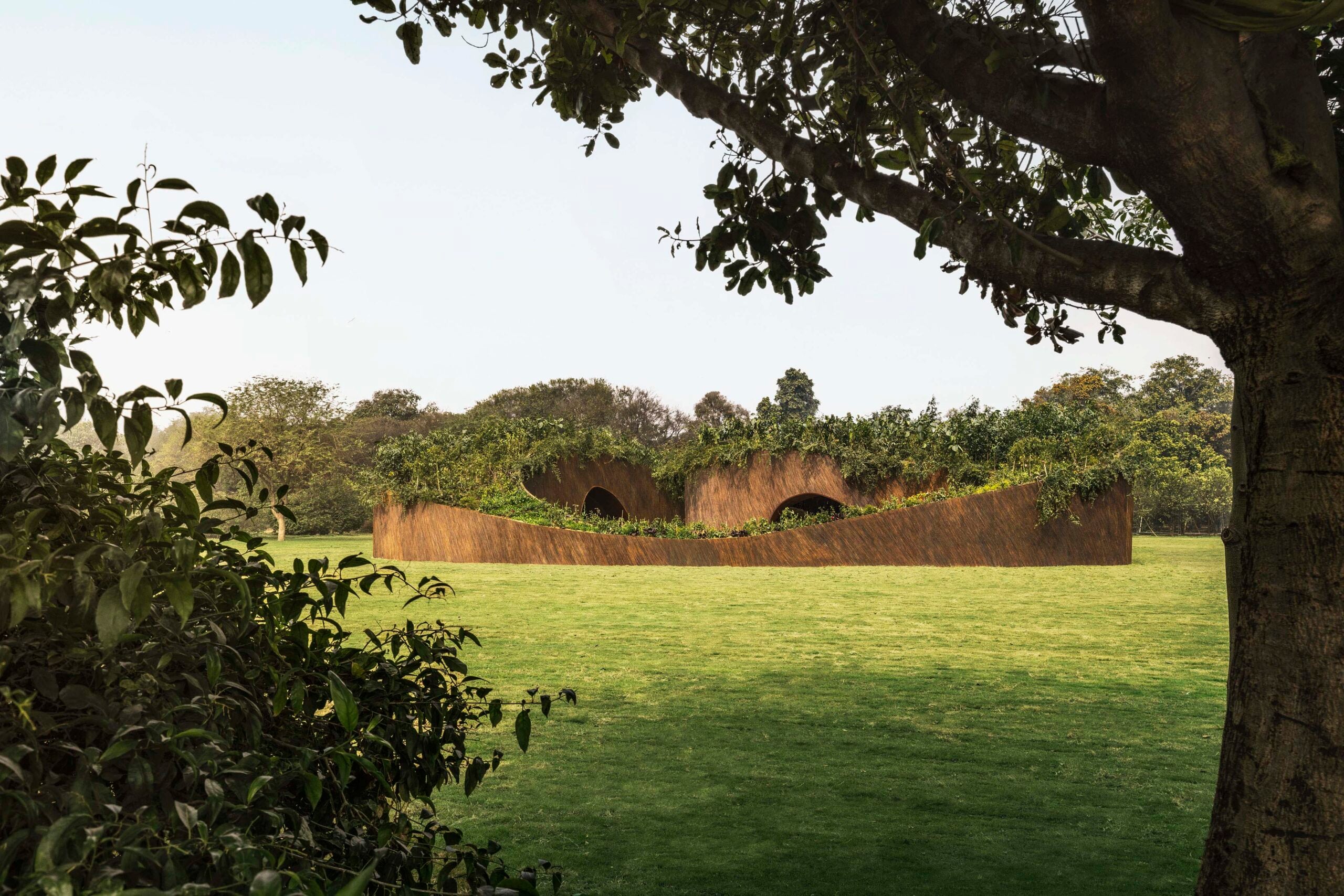 A wide shot of the pavilion from a distance, showing it as a low-lying, undulating structure that blends into the flat green lawn under the shade of a large tree.