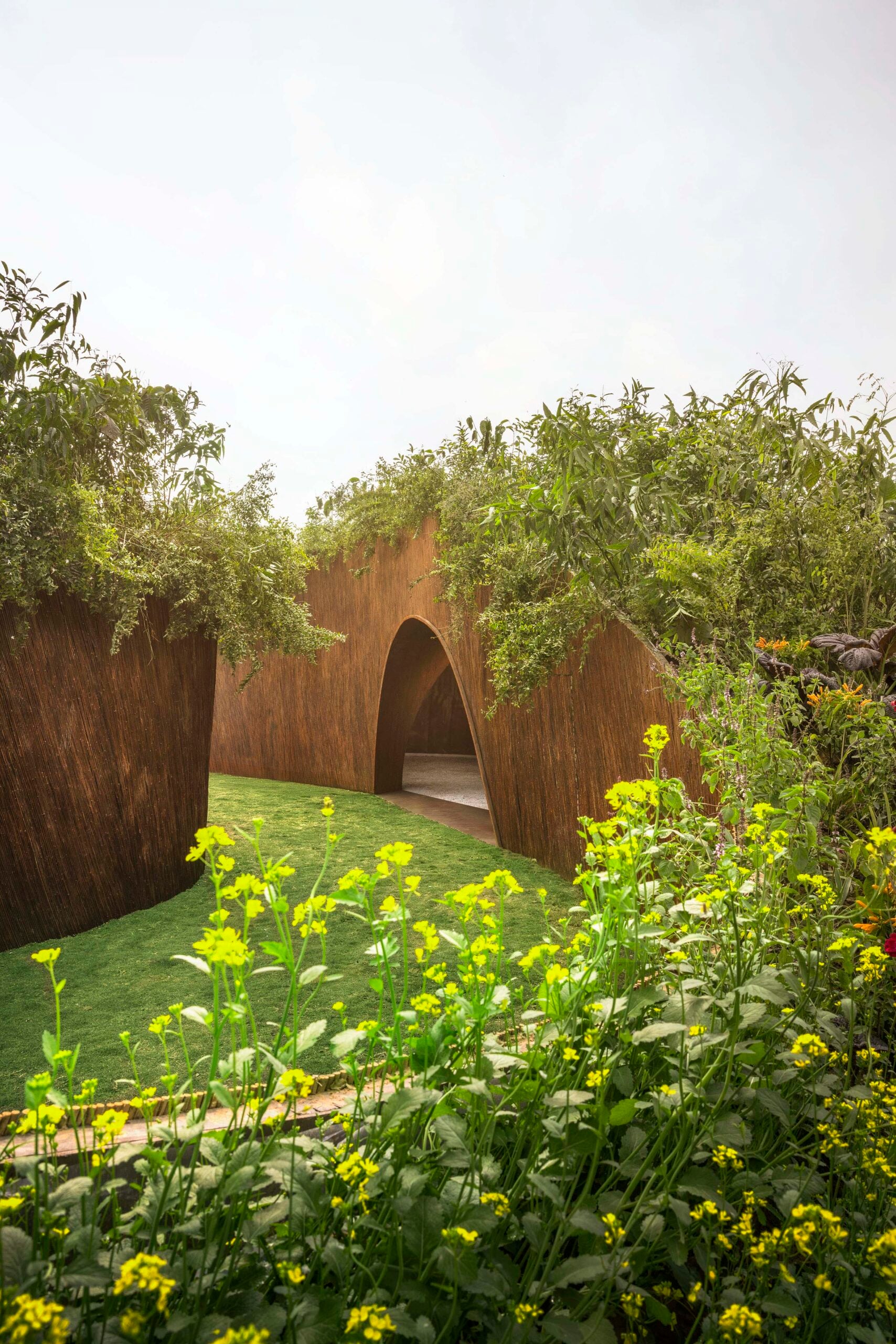 A ground-level view of the pavilion's exterior, showing a thick, textured earthy wall topped with climbing green plants. Bright yellow wildflowers are in focus in the foreground.