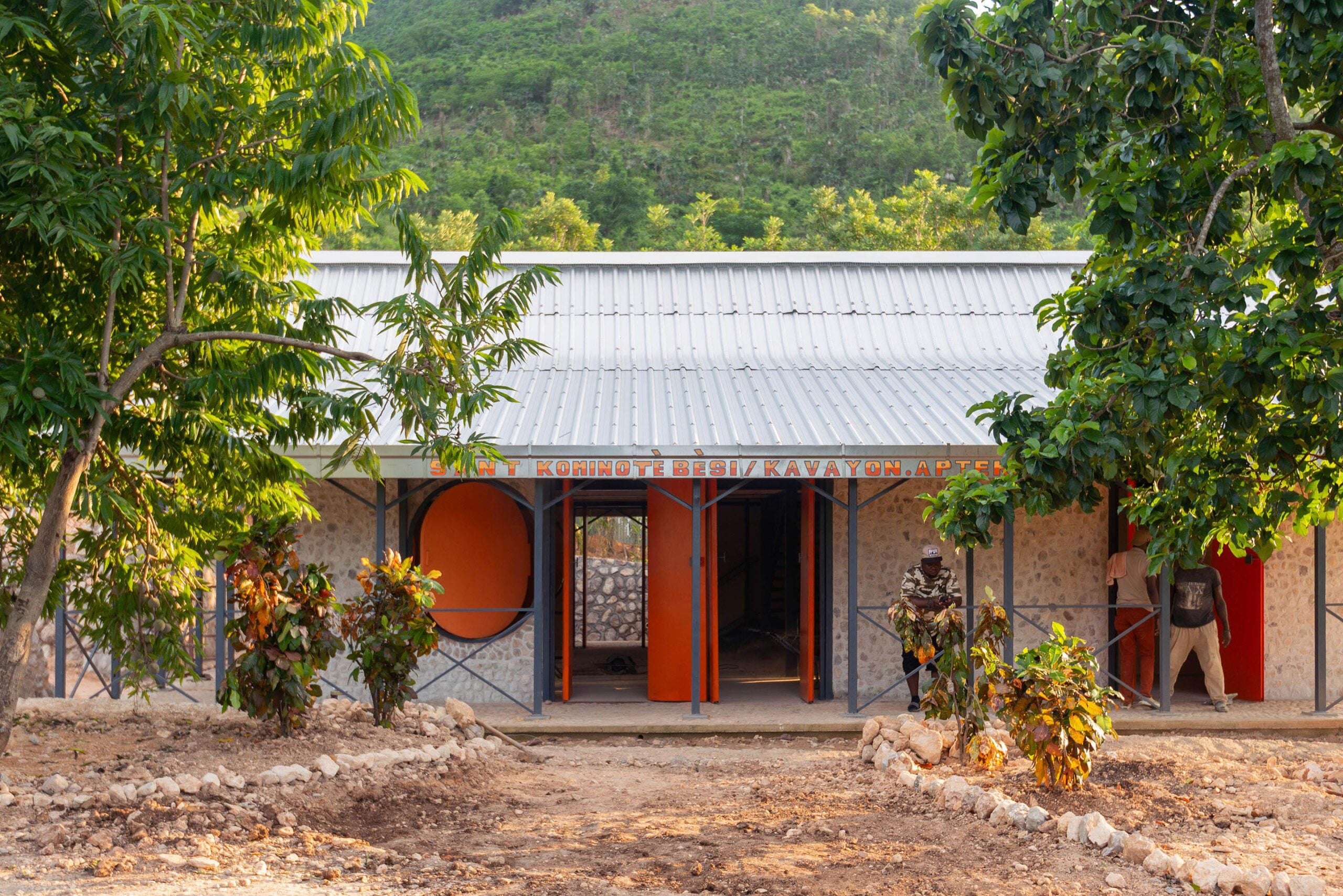 A perspective shot of a single-story community building featuring a large, silver corrugated metal gable roof and textured earth-toned walls. The facade is punctuated by distinctive large, bright orange circular shutters. The building is surrounded by young tropical plants and trees under a cloudy sky.