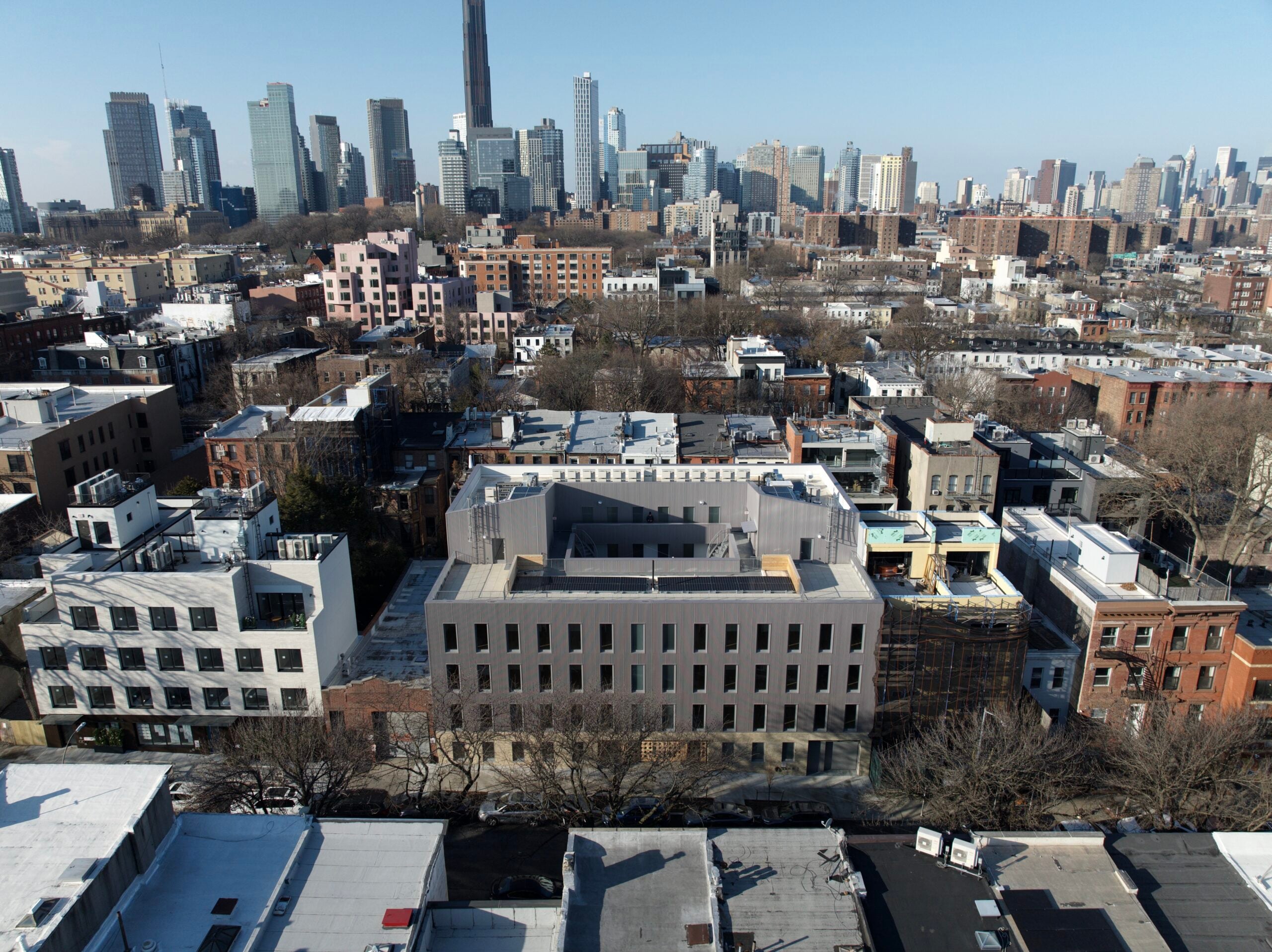 An aerial drone shot of the rectangular mid-rise building showing its central open courtyard. The building is situated in a dense urban neighborhood with a city skyline visible in the distance.