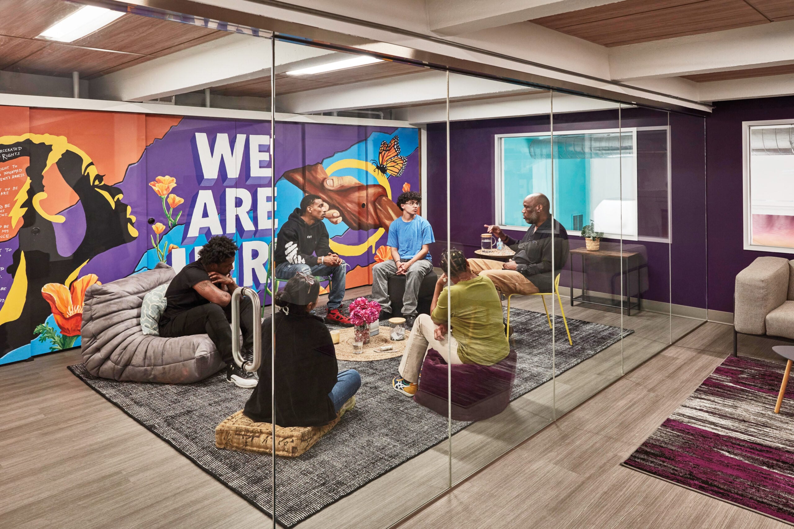 A group of six people sit in a circle on poufs (one sits in a grey Togo) in a glass-enclosed meeting room, with purple walls and a colorful mural.
