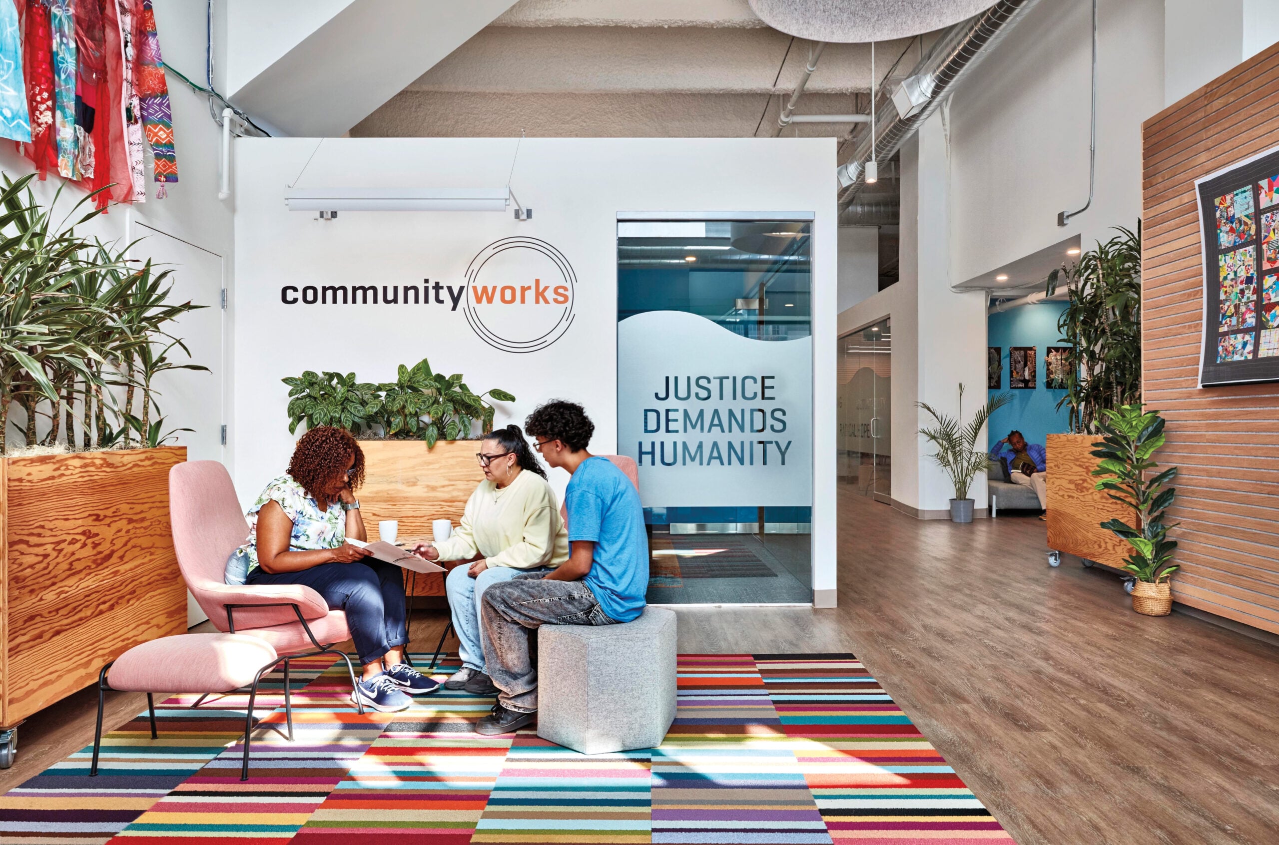Three people sitting in chairs in a seating area. A sign in the background reads: Community Works and Justice Demands Humanity.