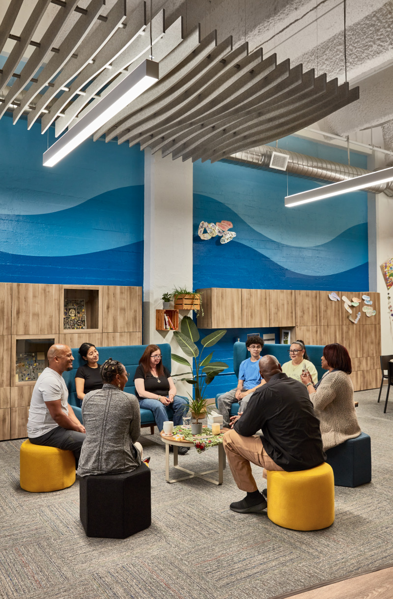 Office interior with exposed ductwork, a blue wave mural, wooden cabinetry and a group of people sitting in a circle