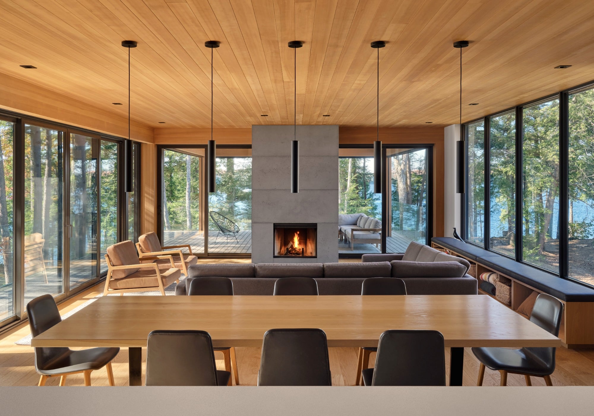 Interior cottage living space with stone fireplace and floor-to-ceiling glass windows and sliding doors, revealing forest outside