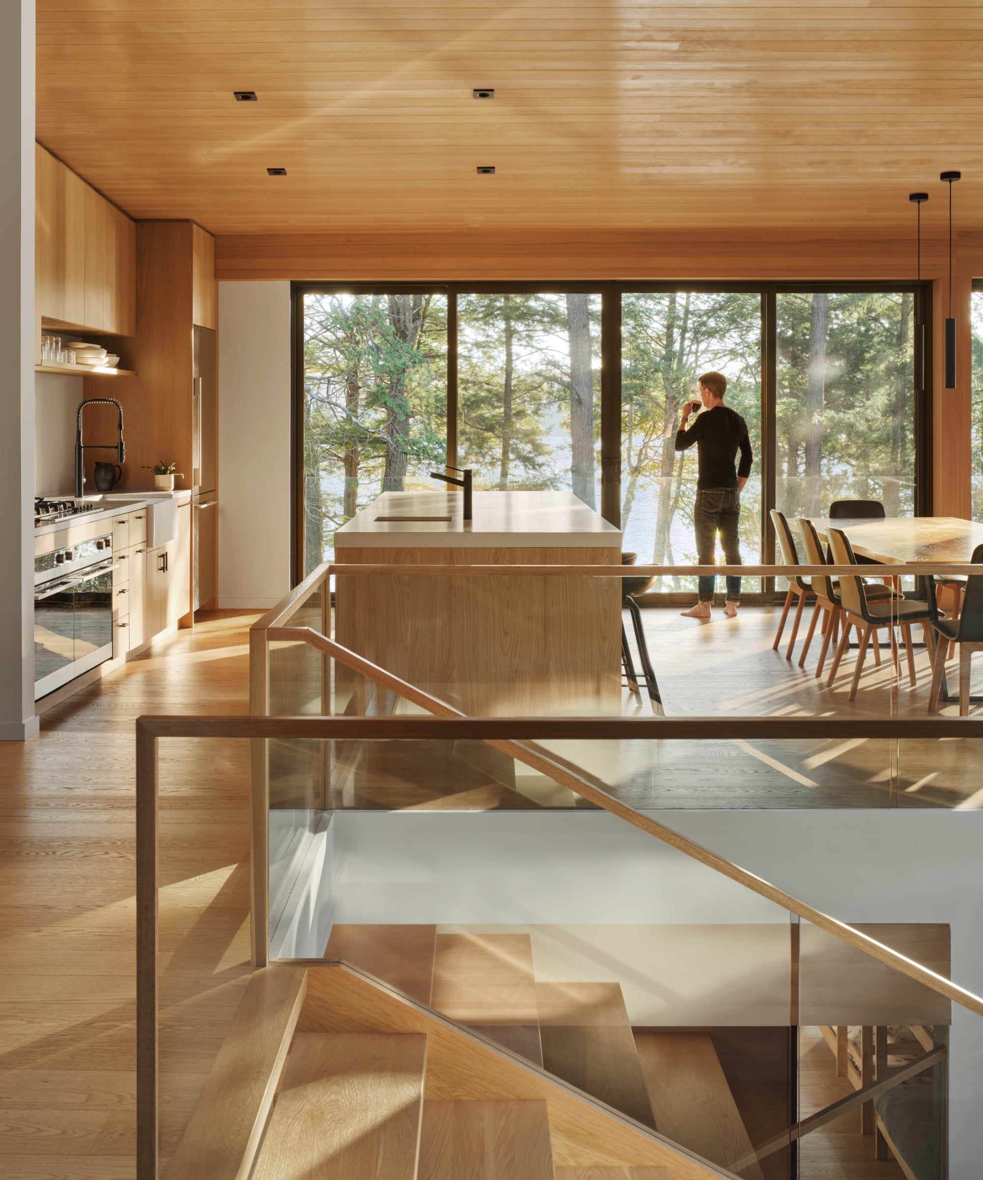 Wood cottage interior with glass floor-to-ceiling windows and a figure sipping coffee.