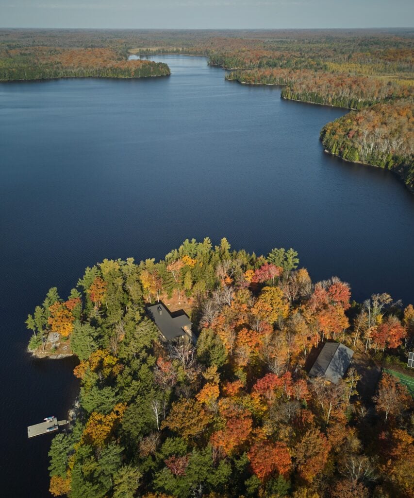 Birds-eye view of a peninsula in Muskoka Lake with leafy trees in autumn colours, blue water and the roof of a cottage.