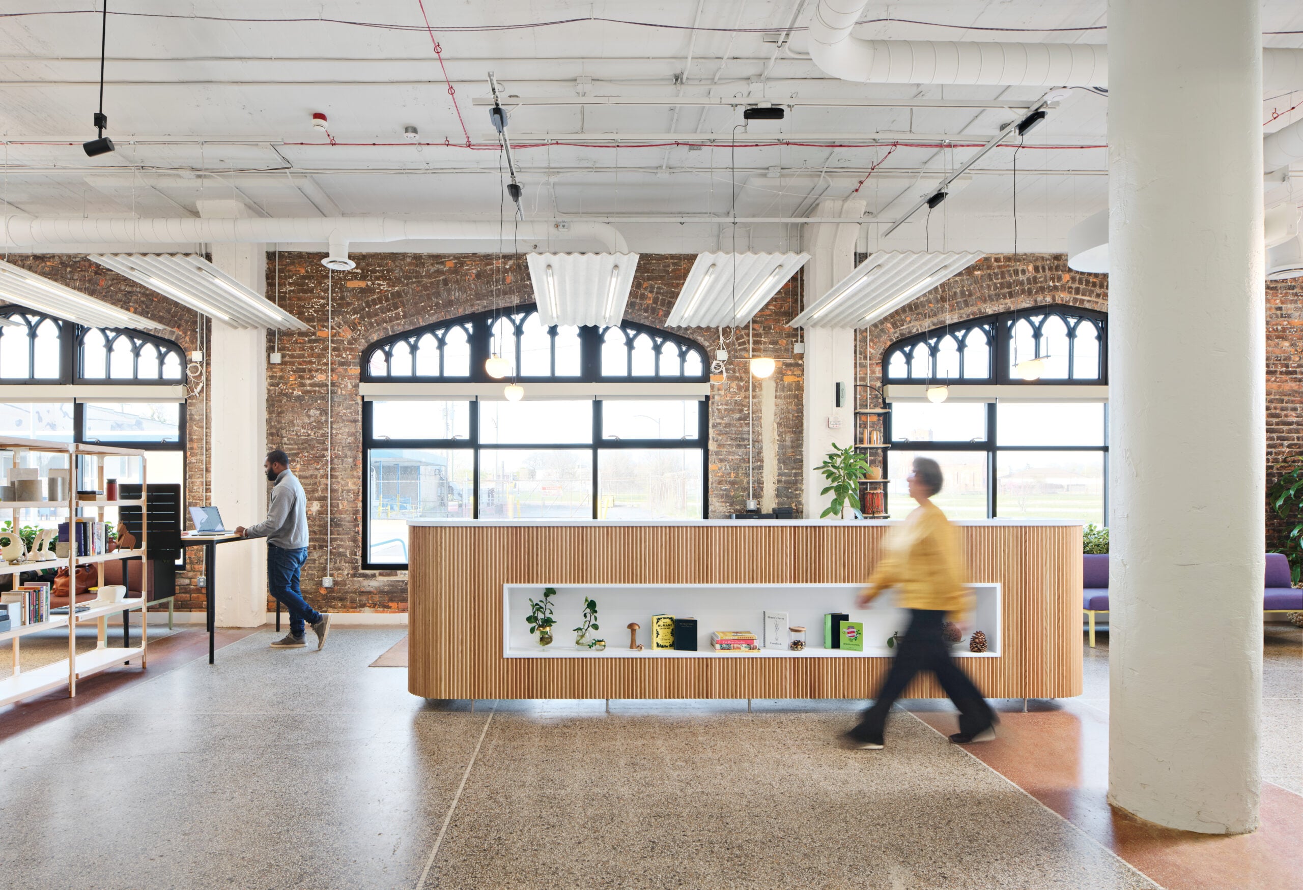 LOVE Building interior, featuring large bright windows, exposed brick, concrete pillars, and modern furnishings with two figures.