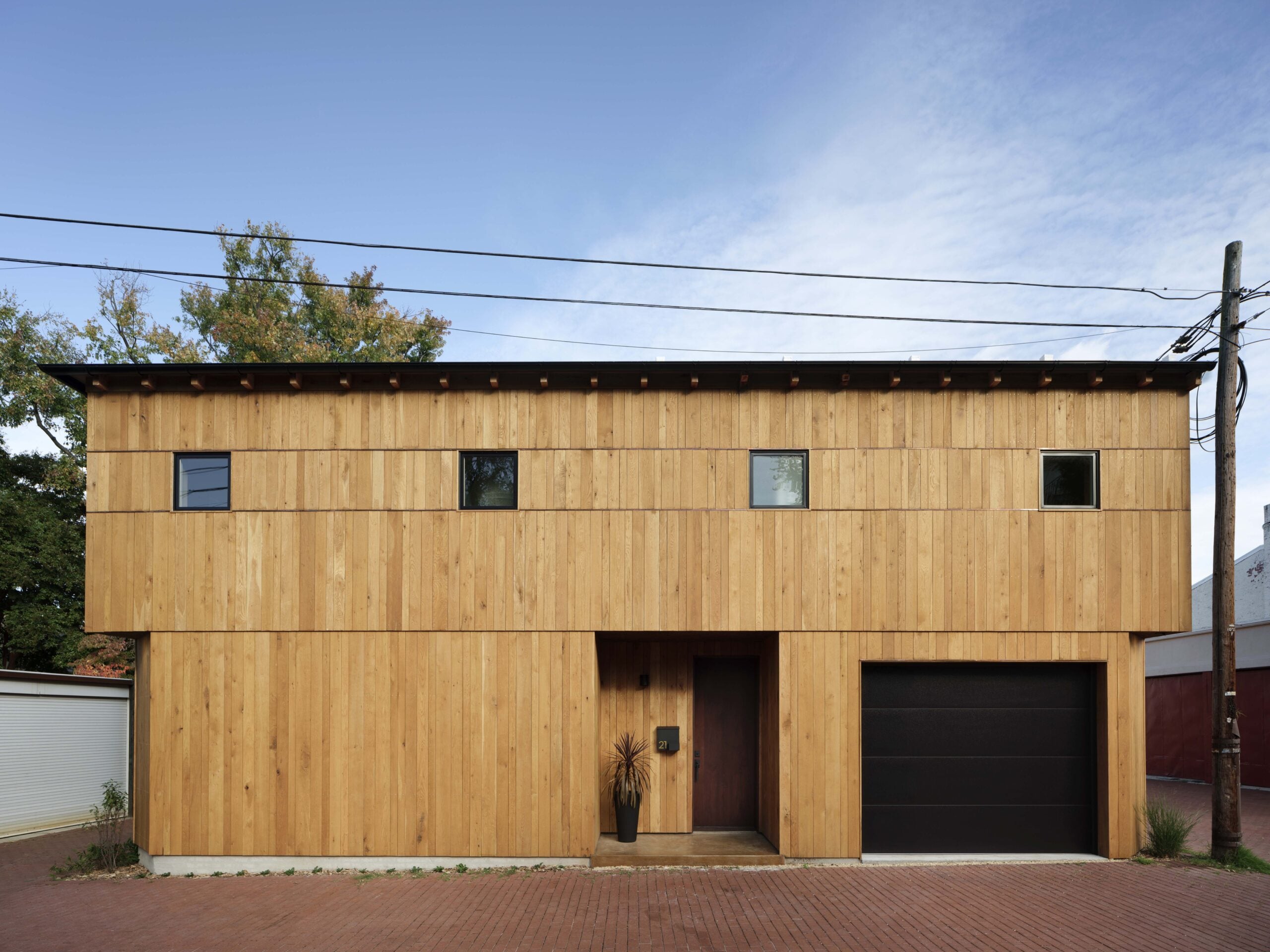 A modern two-story house with a flat roof, wooden exterior, small square windows, a black garage door, and a potted plant by the entrance.