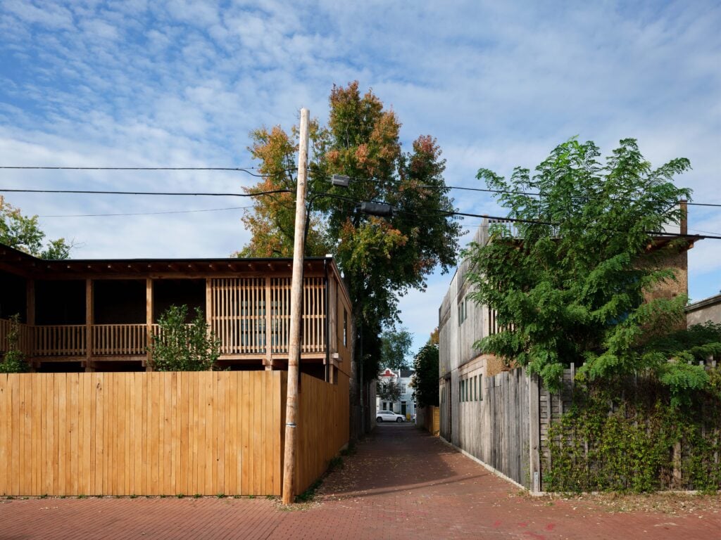 A narrow alleyway runs between a wooden-fenced building on the left and a gray building on the right, with trees and power lines visible under a partly cloudy sky.