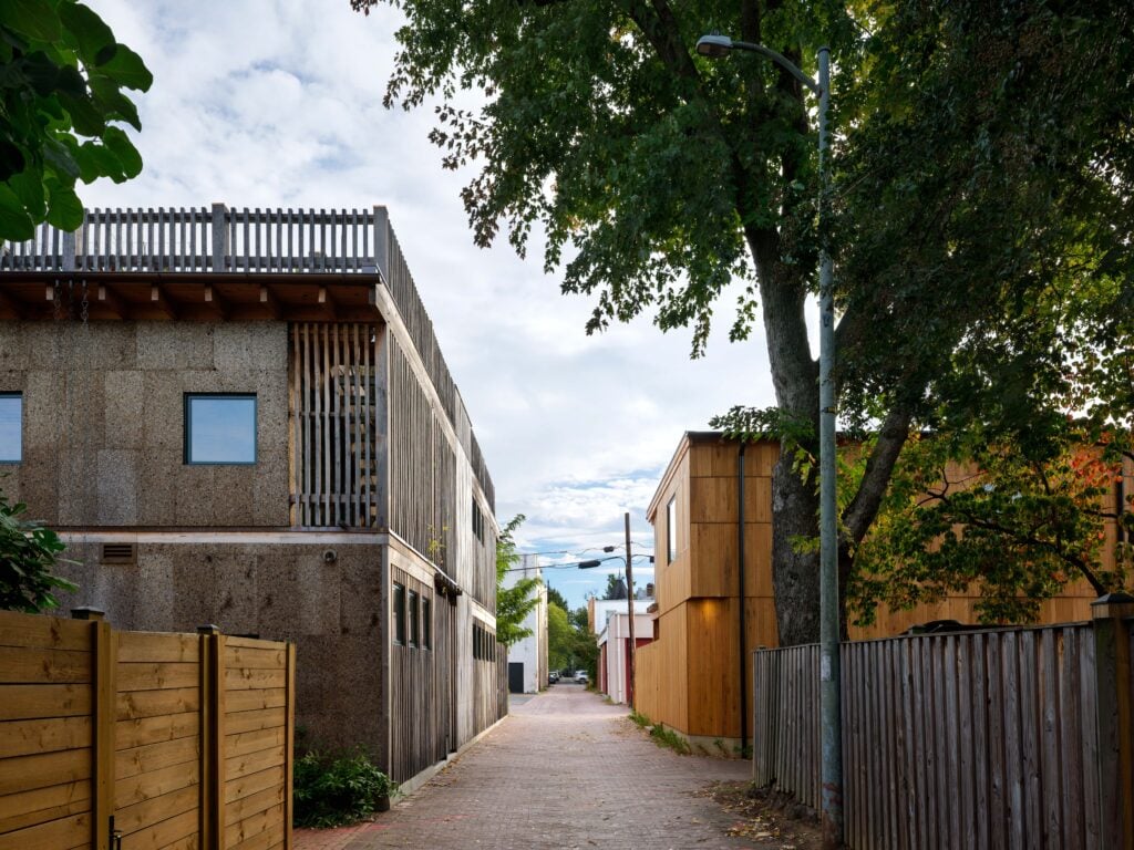 A narrow alleyway lined with modern wooden and concrete houses, tall trees, wooden fences, and overhead utility wires under a partly cloudy sky.