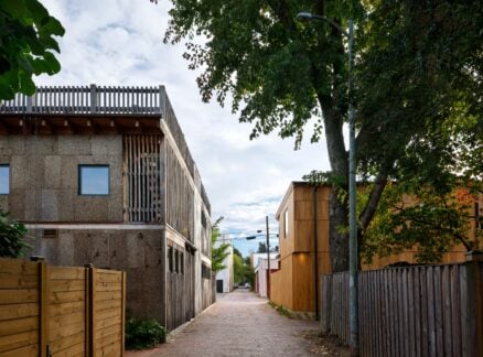 A narrow alleyway lined with modern wooden and concrete houses, tall trees, wooden fences, and overhead utility wires under a partly cloudy sky.
