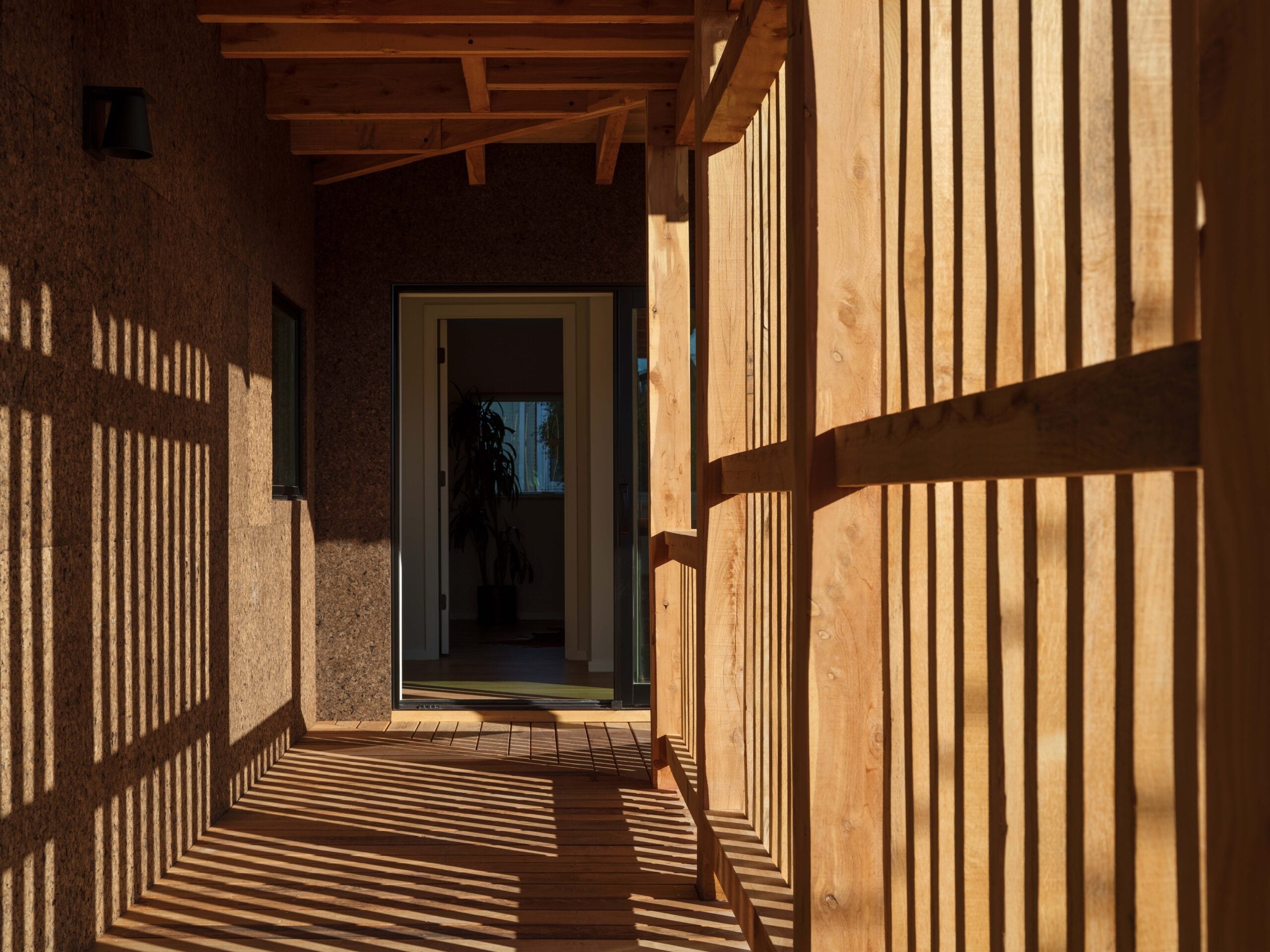 A wooden corridor with vertical slats casting shadows, leading to an open doorway with a glimpse of an indoor plant and sunlight beyond.