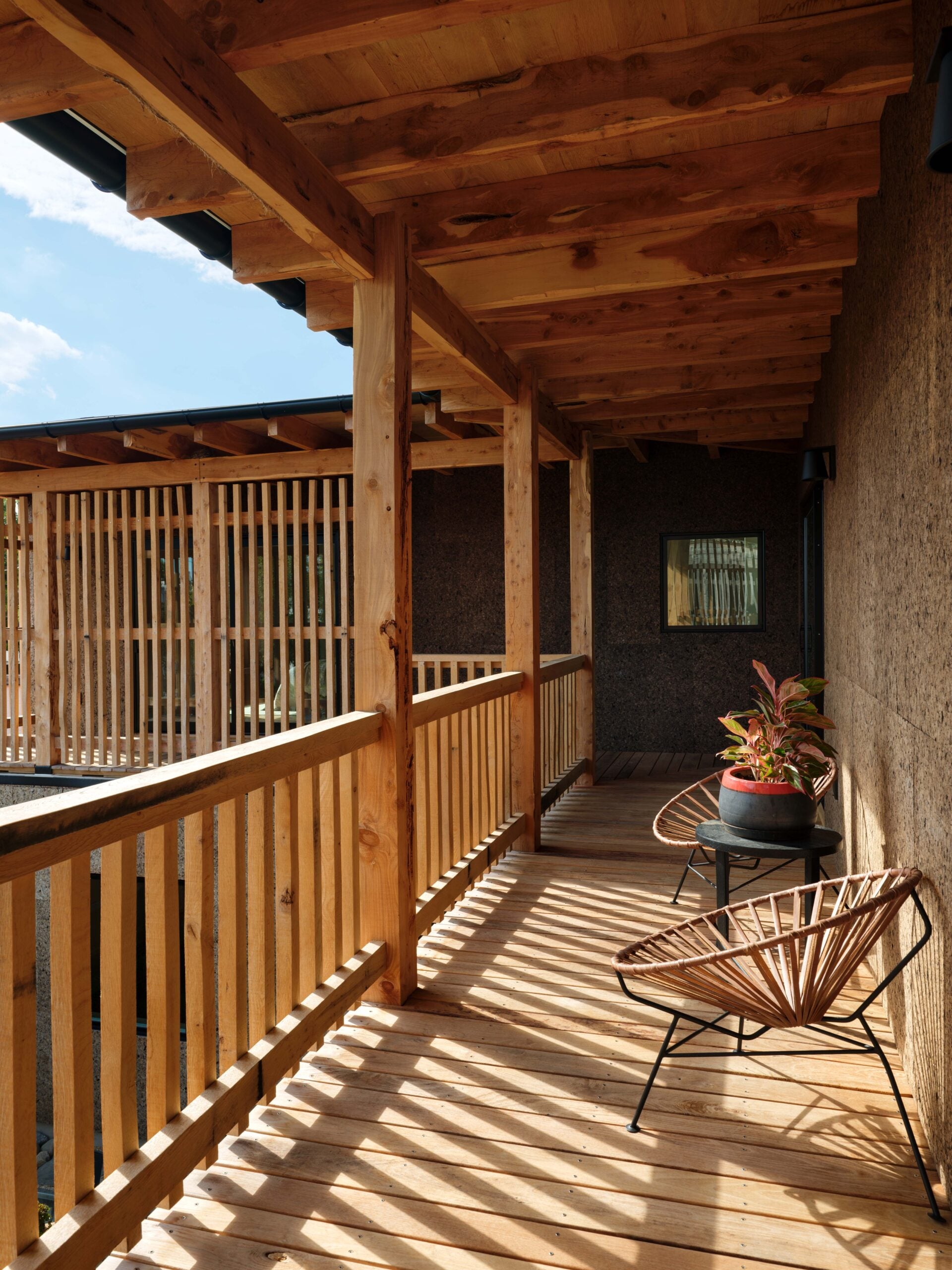 Wooden balcony with slatted railing, two modern chairs, a potted plant, and sunlight casting shadows across the floor.