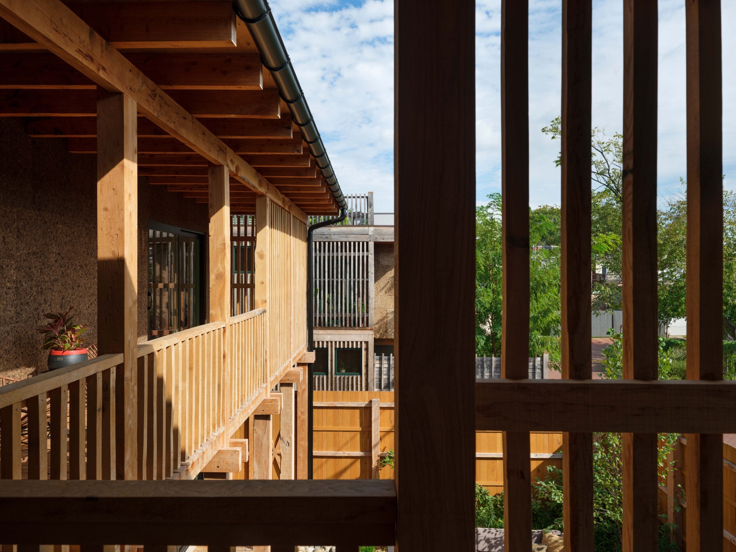 View of a wooden building exterior with vertical slats, open corridors, and a potted plant on the railing, set against a backdrop of trees and a partly cloudy sky.