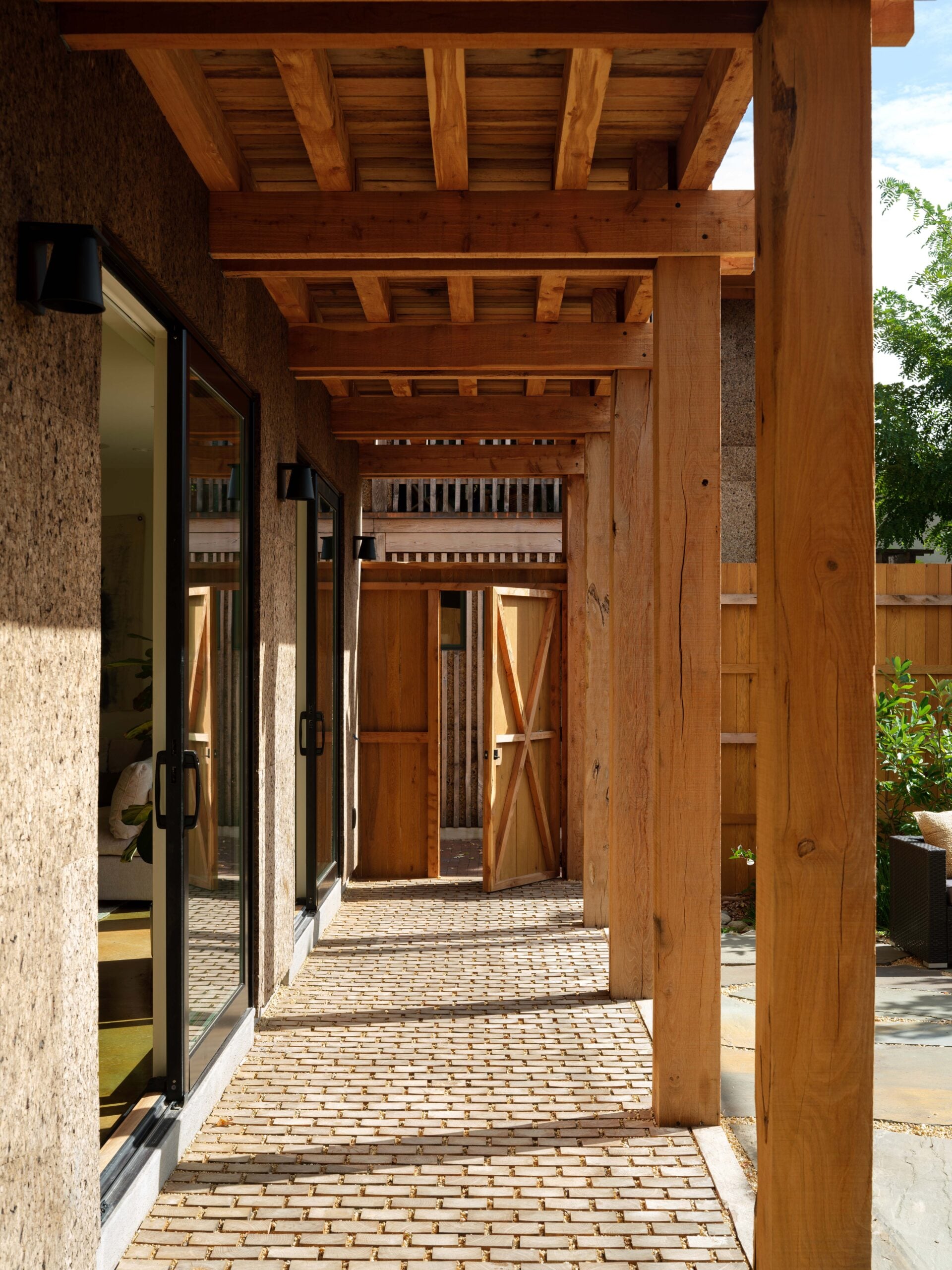 Covered walkway with wooden beams and brick flooring, sliding glass doors on the left, and a wooden gate at the end, lit by natural sunlight.