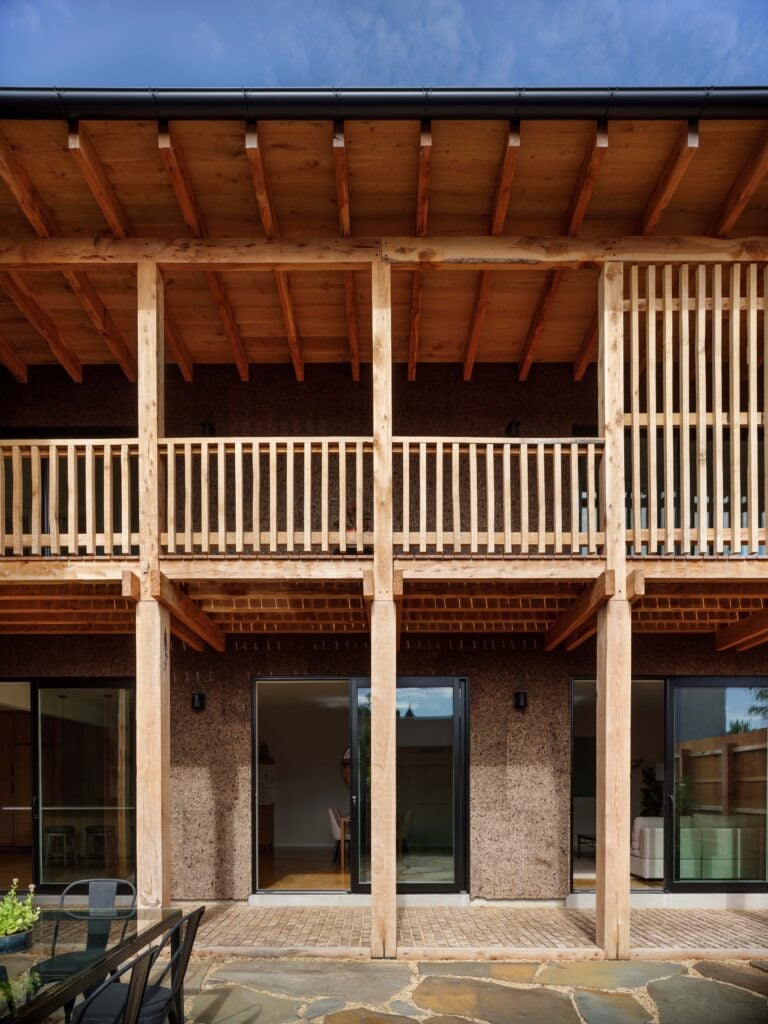 Two-story building with wooden beams, railings, and columns; exterior features sliding glass doors and a textured brown wall under a sloped roof.