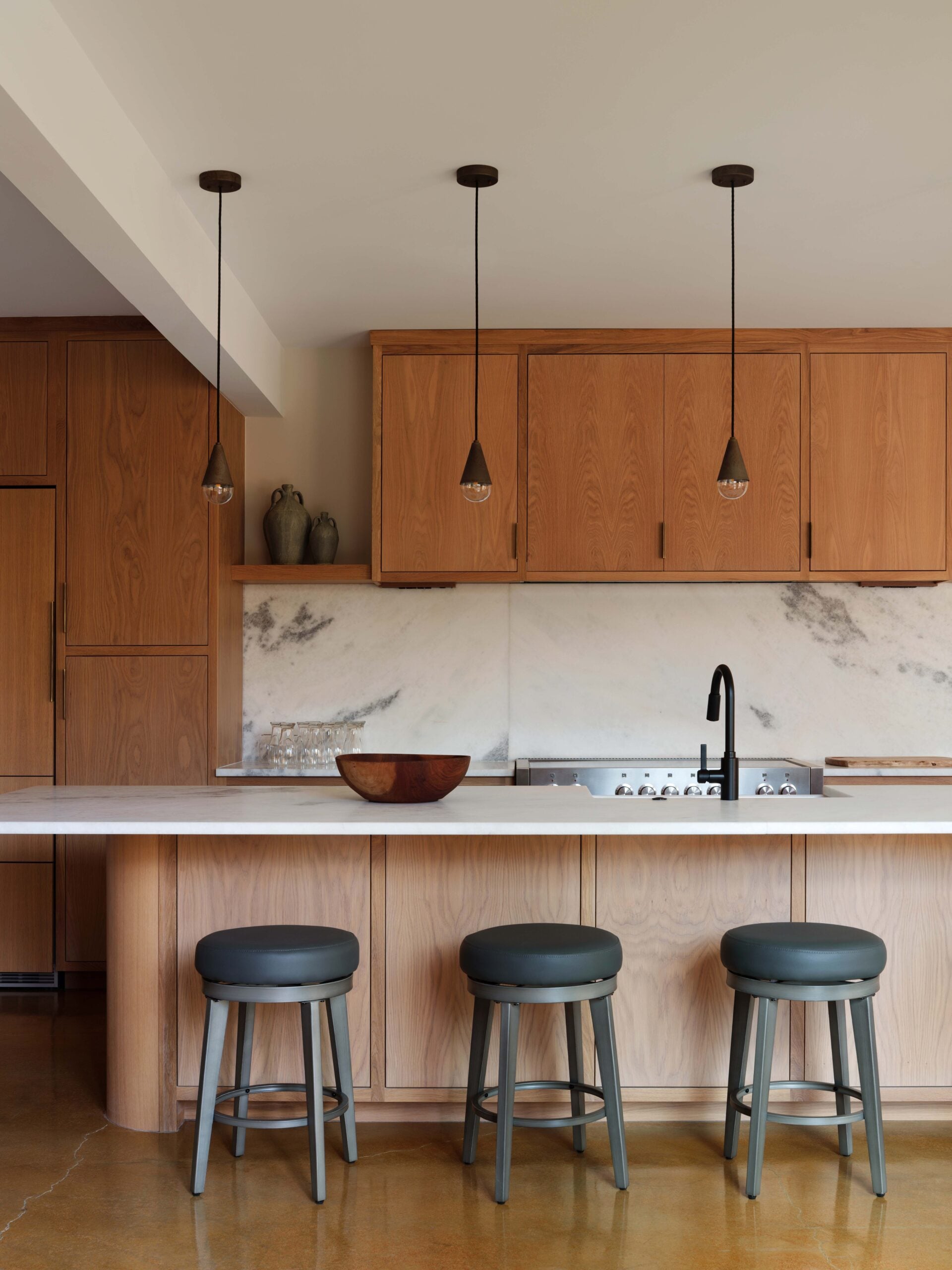 Modern kitchen with wood cabinets, marble backsplash, three gray stools at an island, pendant lights above, and a bowl on the countertop.
