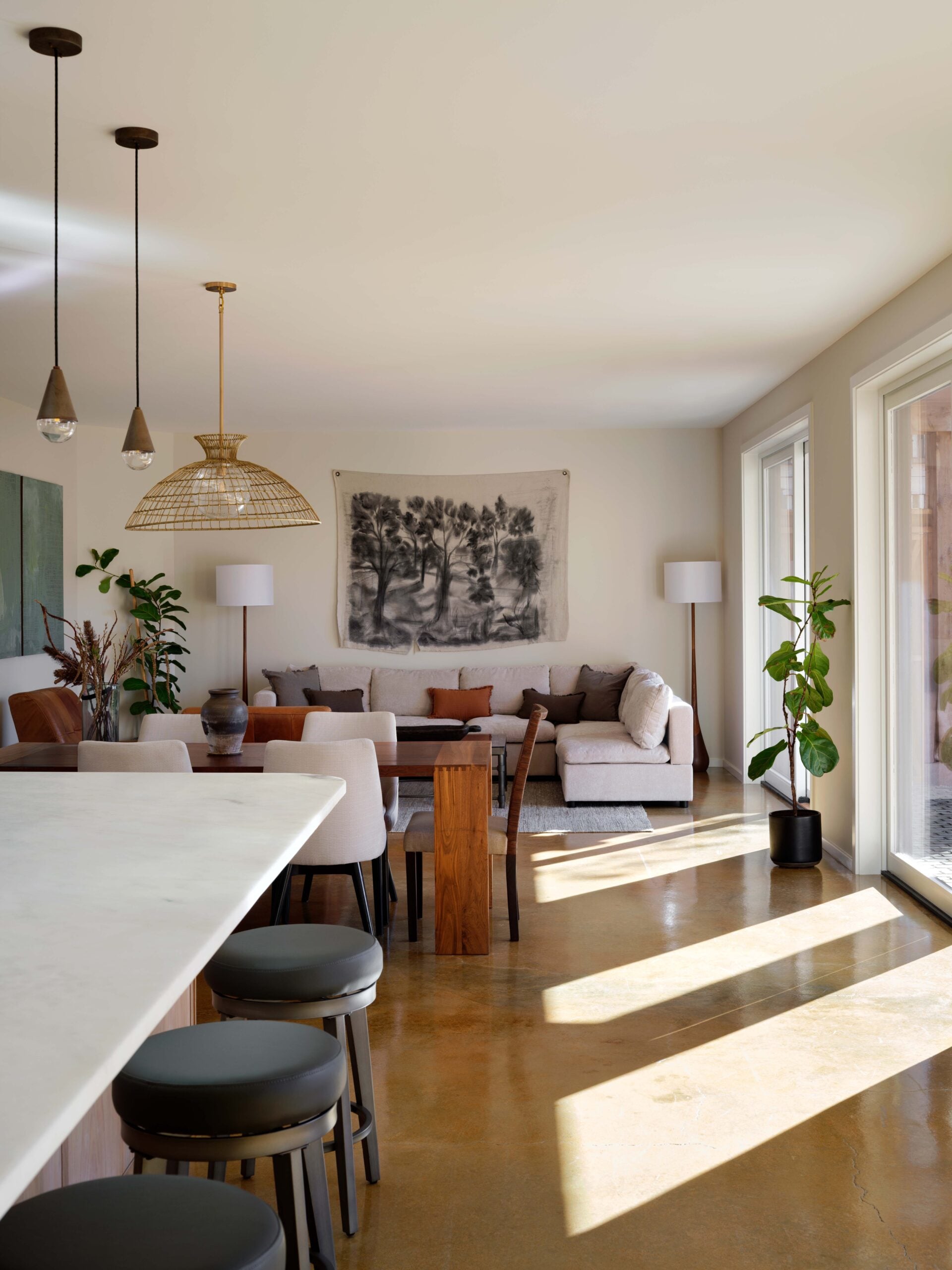 Contemporary open-plan living and dining area with neutral tones, modern furniture, potted plants, large windows, and artwork on the wall. Sunlight streams across polished floors.