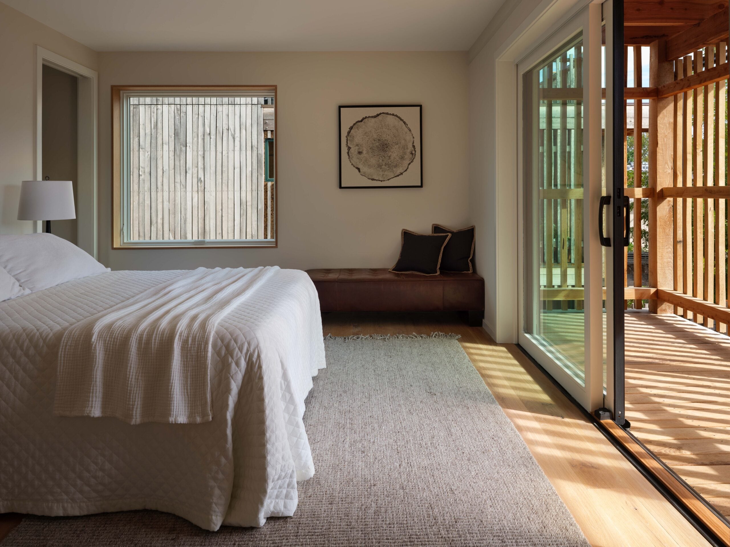 A minimalist bedroom with a bed, a bench under a window, a framed artwork, and sliding glass doors opening to a wooden balcony.