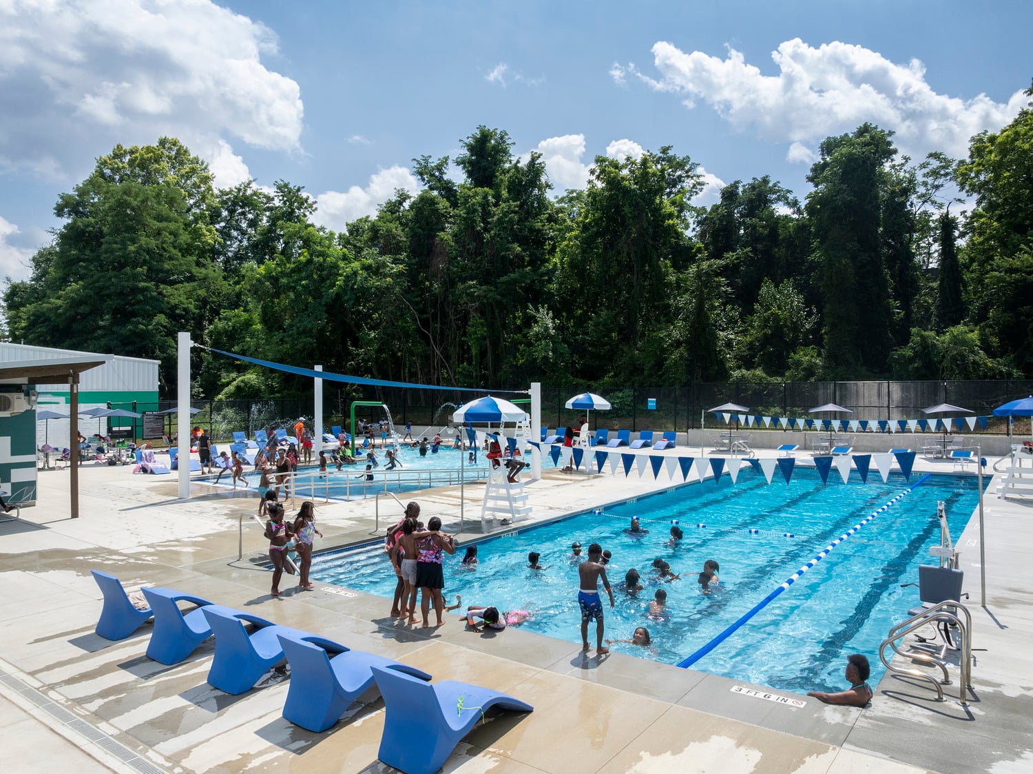 Outdoor pool with blue lounge chairs, bright blue water and large green trees in the background, full of people swimming