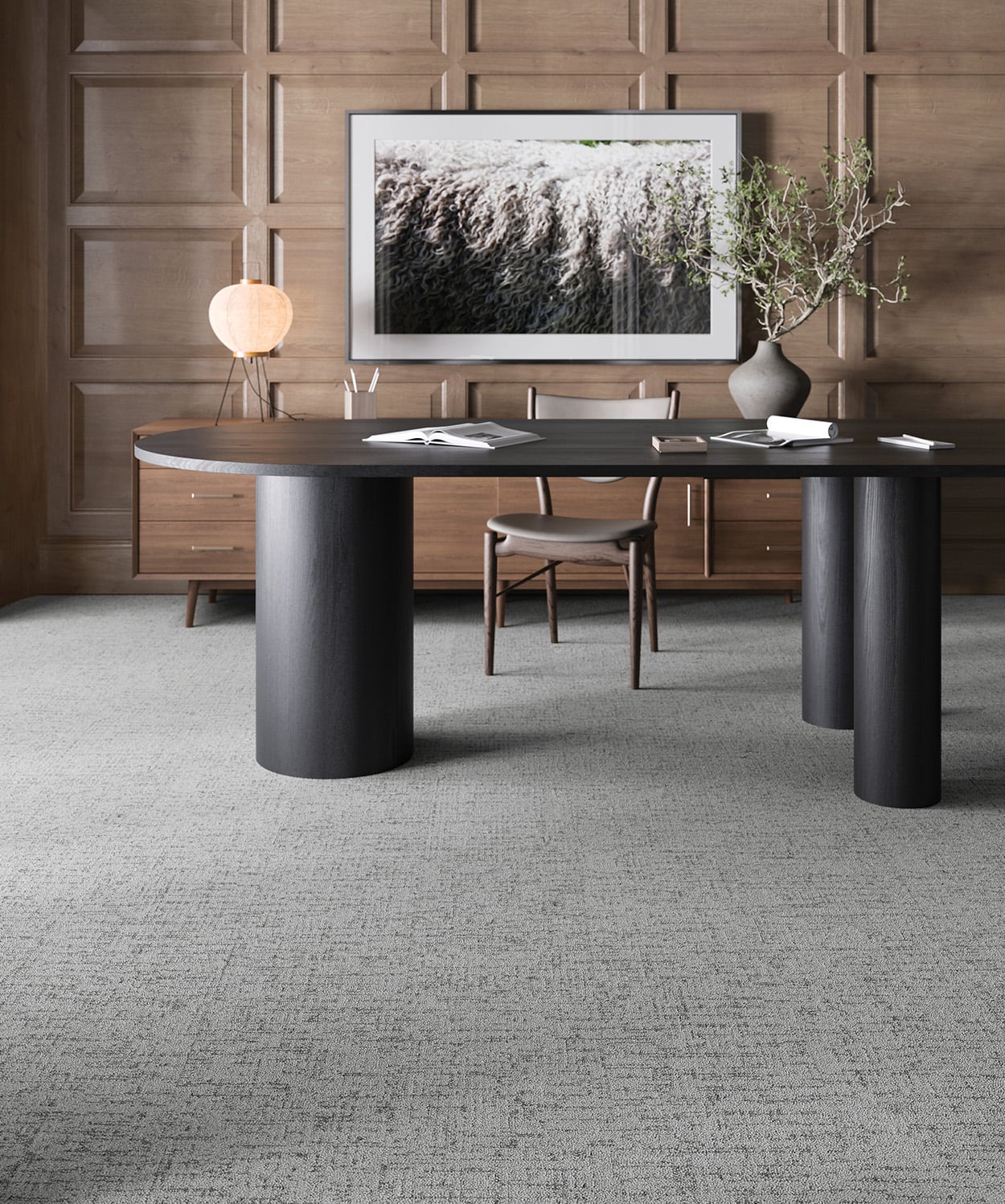 A modern office with a round black table, wooden chair, sideboard, lamp, framed wall art, and a potted branch, set against wood-paneled walls and gray carpet.