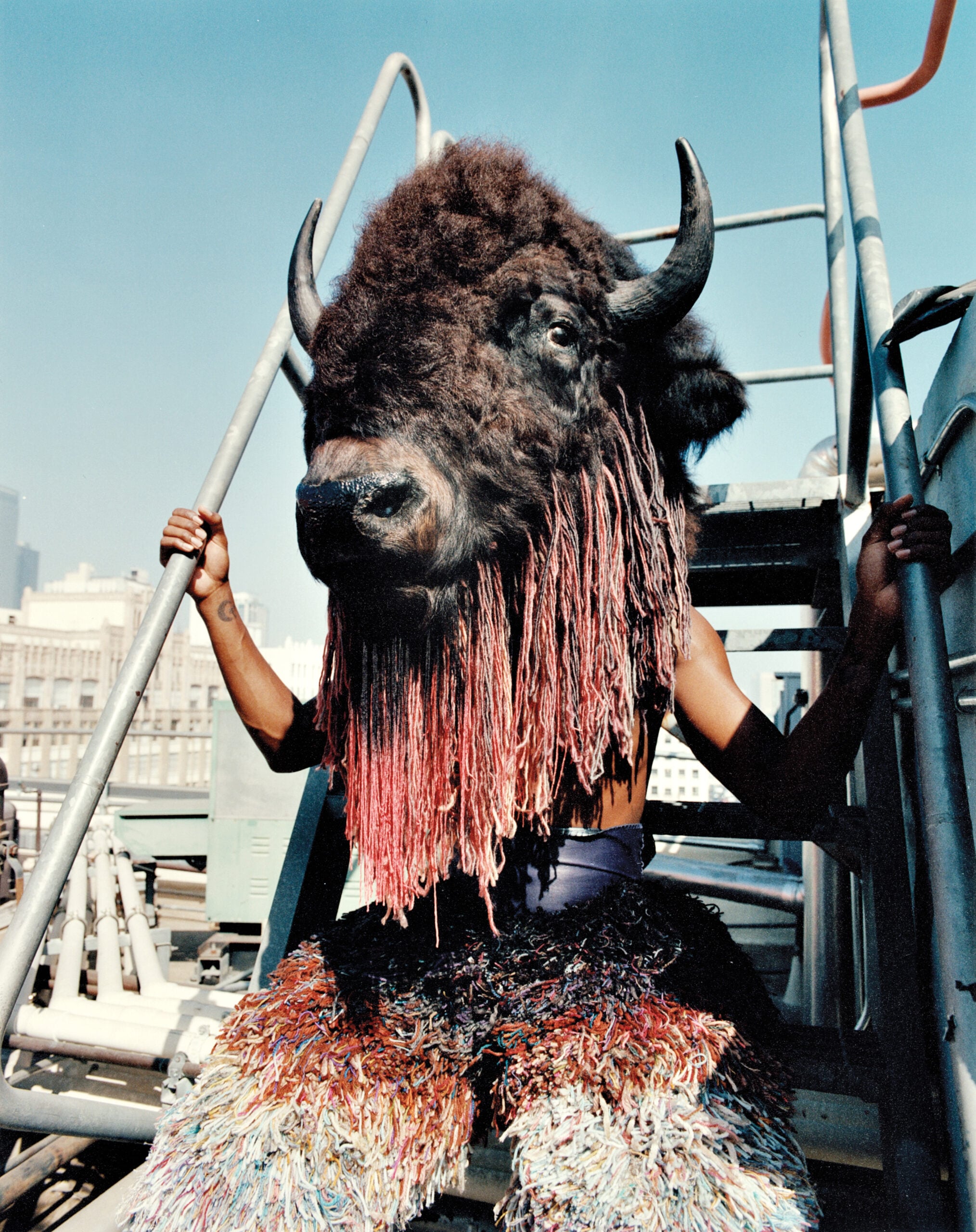 Person wearing a large bison head and tufted pants sits on a fire escape with blue sky background