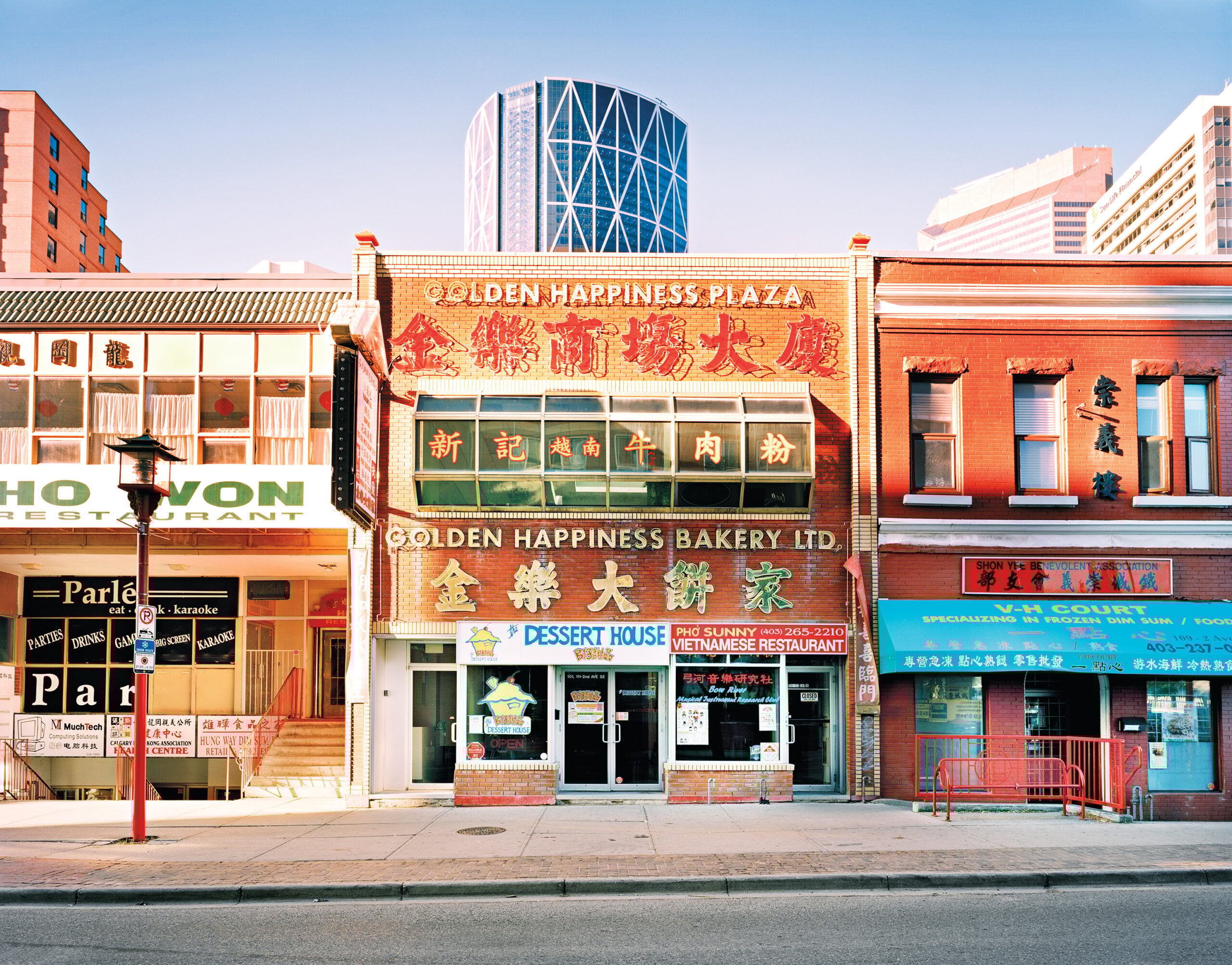 Street view of a row of low-rise brick buildings in a Chinatown district under bright daylight. The central building features large signage reading “Golden Happiness Plaza” and “Golden Happiness Bakery Ltd.” in English and Chinese characters, with red and gold lettering.
