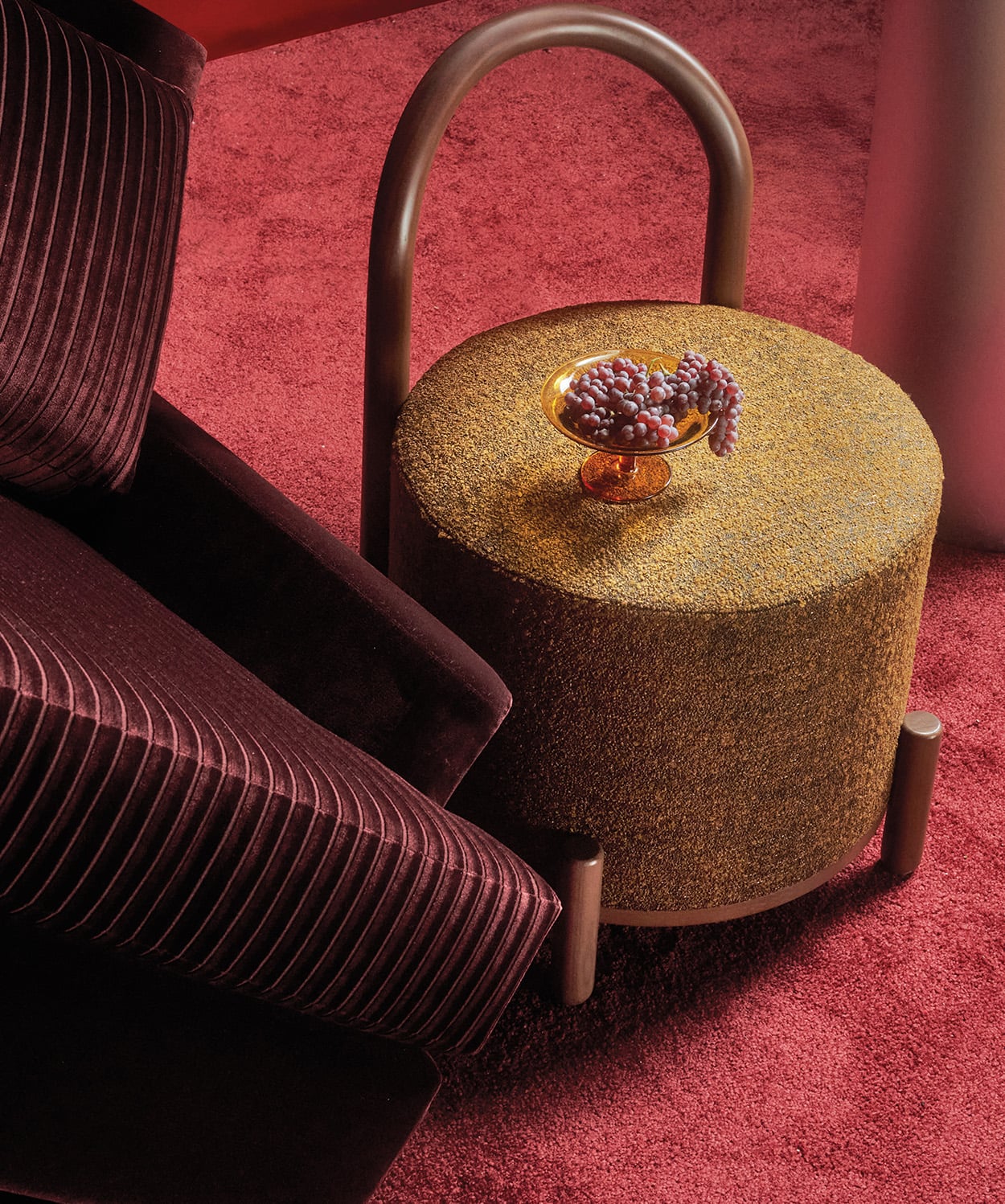 A gold textured round side table with a curved handle holds a small bowl of grapes, next to a dark velvet chair on a red carpeted floor.