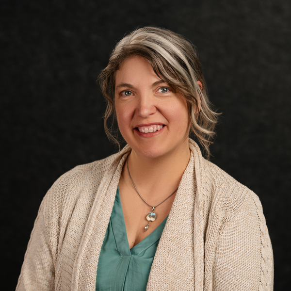 Woman with wavy brown and gray hair wearing a beige sweater and teal blouse, smiling in front of a dark background, celebrating her recognition at the Planet Positive Awards.