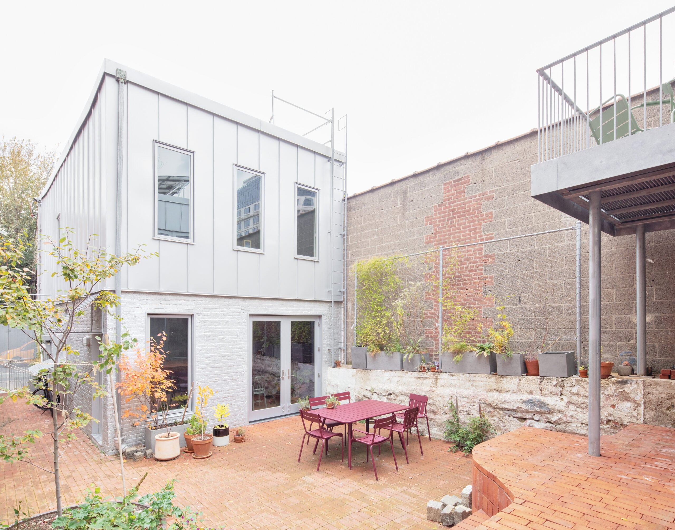 A house with galvanized steel and white brick shares a joint courtyard with another structure, with outdoor seating and potted plants