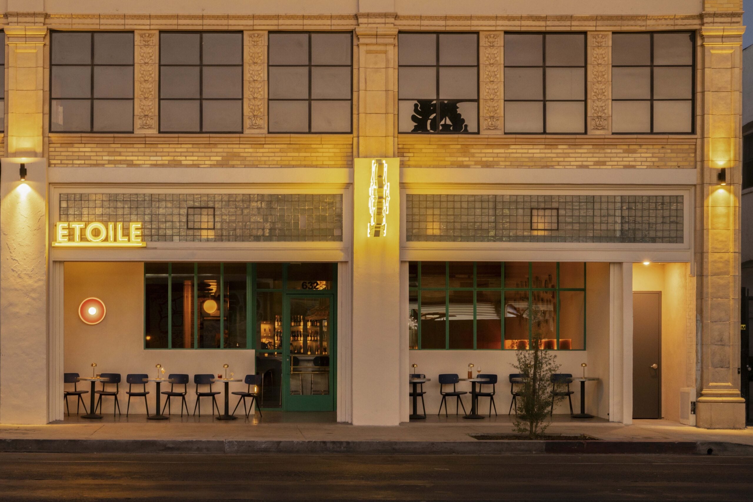 The exterior of a restaurant named "Etoile" at night, with lit signage, large windows, and tables with chairs set up along the sidewalk, offering a charming setting where lovers unite under the city lights.