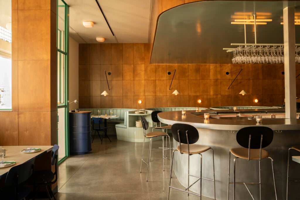 Modern restaurant interior with wooden paneled walls, curved bar with high stools, booth seating, and pendant lighting—an inviting space where lovers unite. Tables are set and wine glasses hang above the bar.