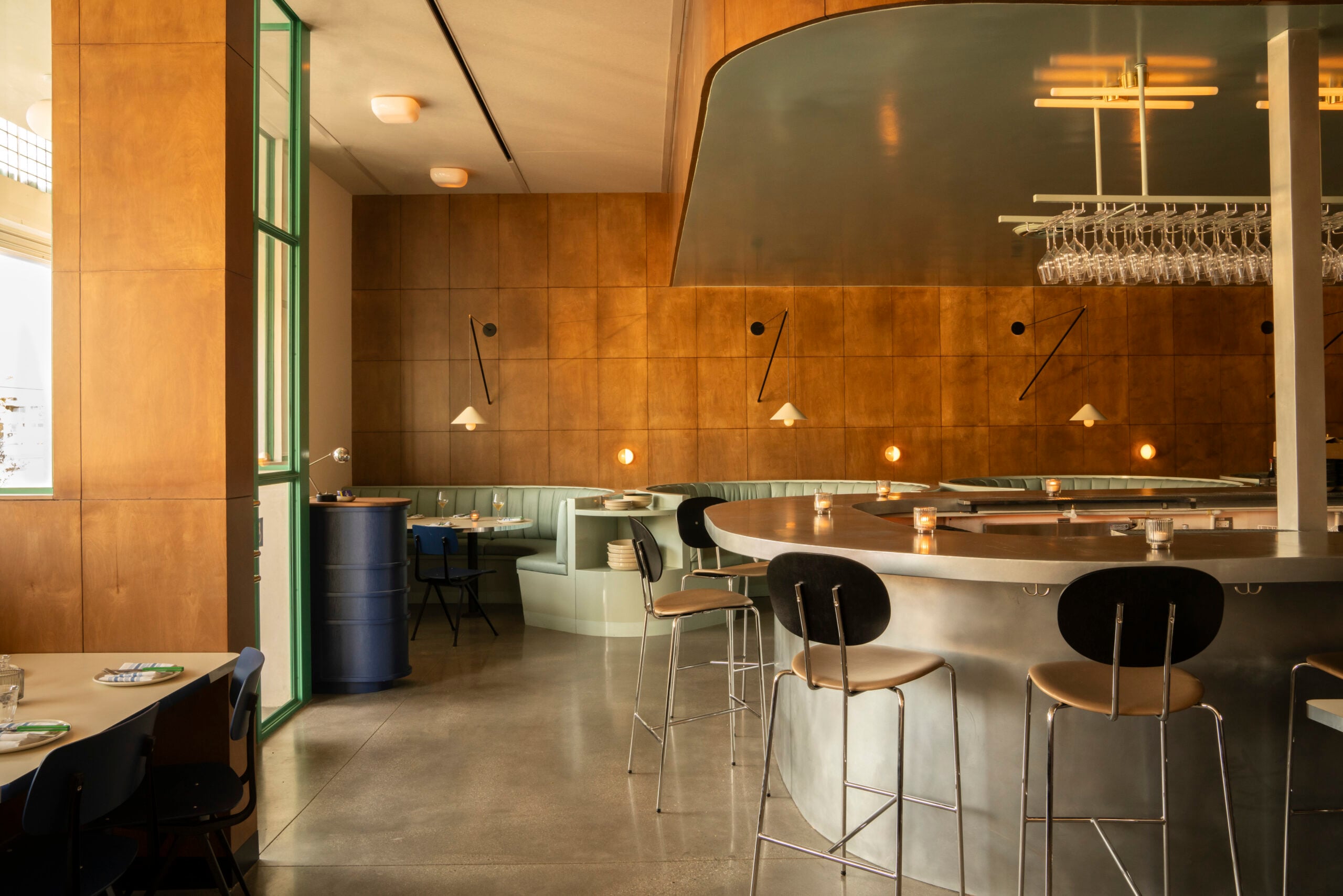 Modern restaurant interior with wooden paneled walls, curved bar with high stools, booth seating, and pendant lighting—an inviting space where lovers unite. Tables are set and wine glasses hang above the bar.