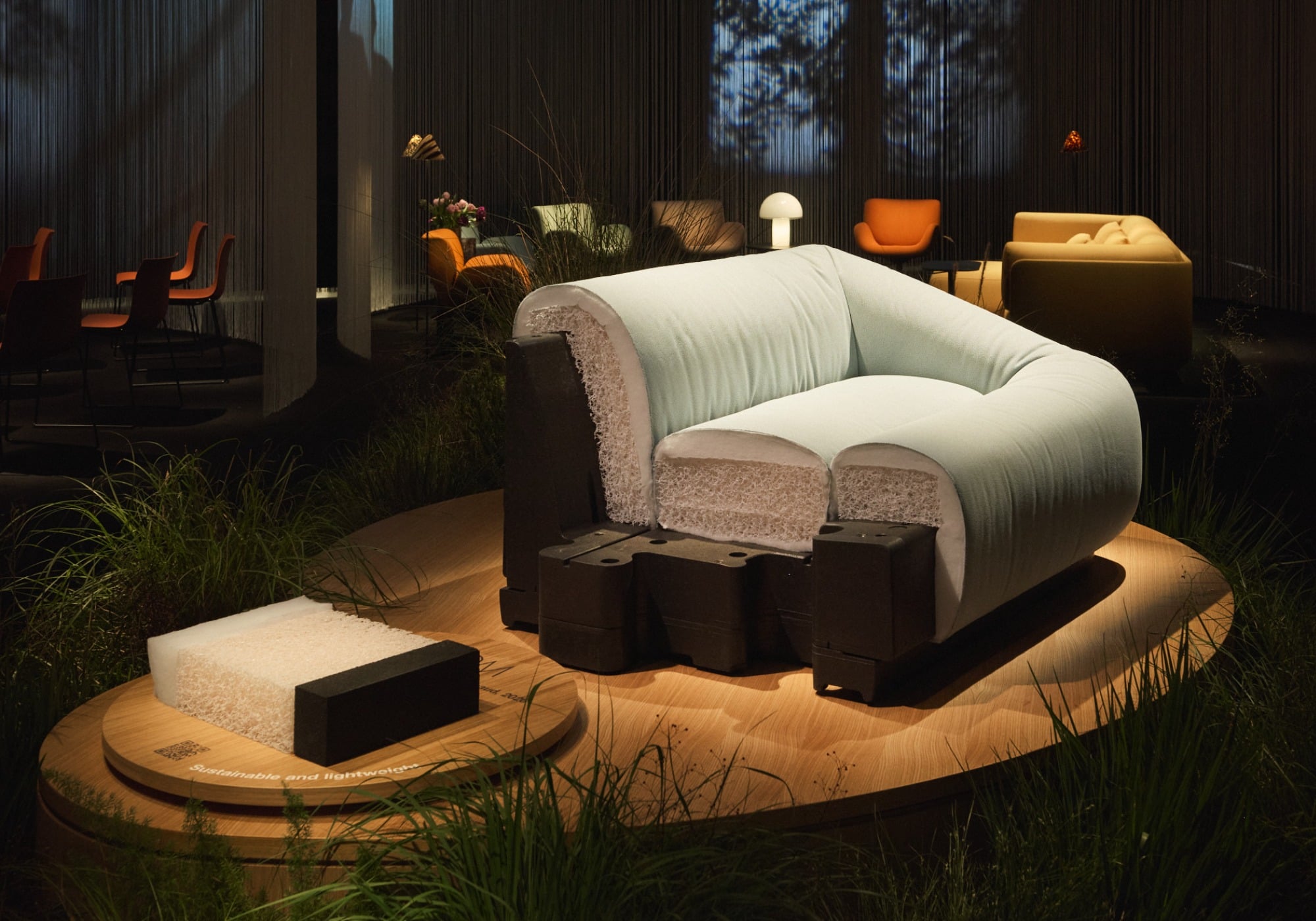 A modern armchair with exposed layers and materials is displayed on a round wooden platform, surrounded by grass and plants—a nod to Milan Design Week’s focus on sustainable products—with various chairs and lighting in the background.